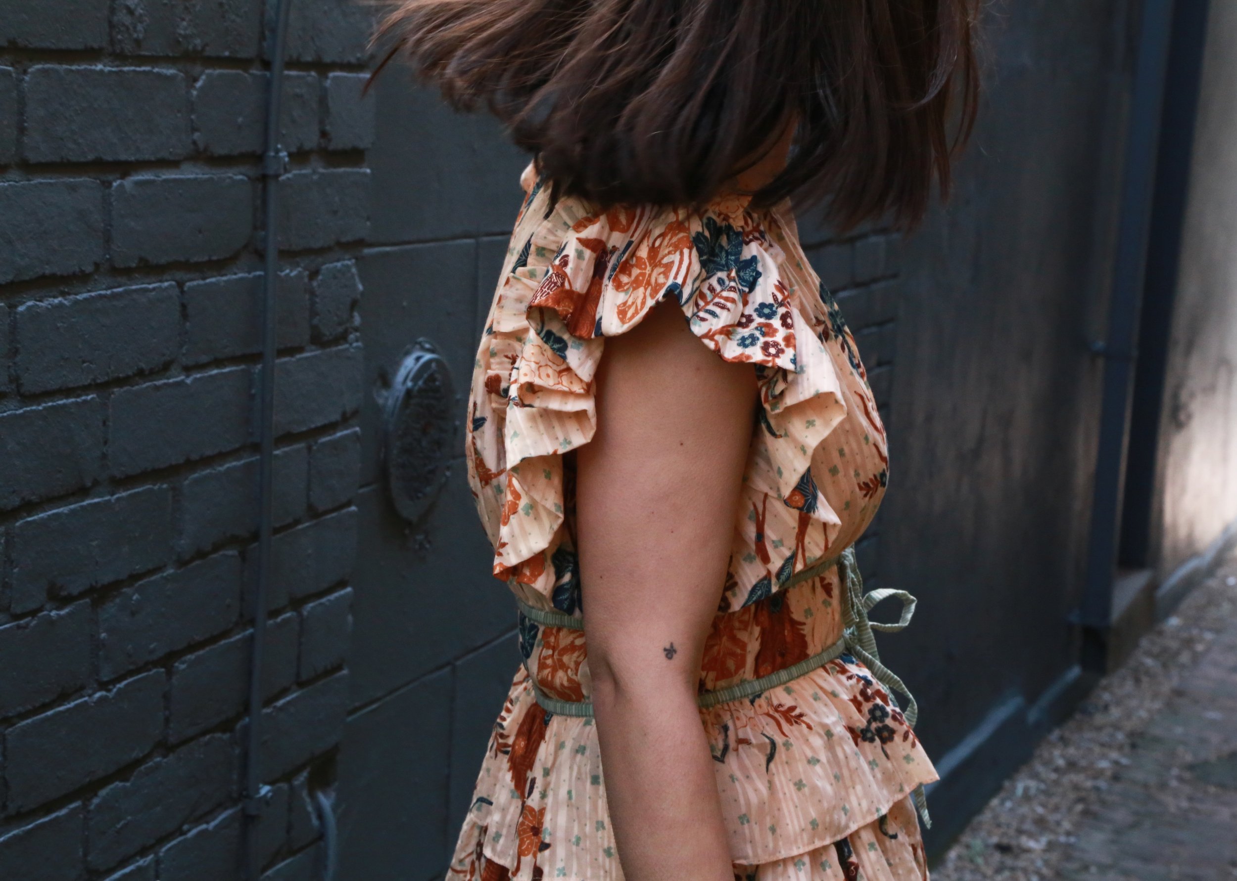 A woman is standing against a dark brick wall, wearing a ruffled floral dress, with her head bowed and her face mostly obscured by her dark hair.