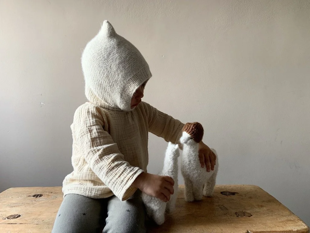 Child in a cozy, hooded sweater and pants petting a small, plush, white sheep figure on a wooden table.