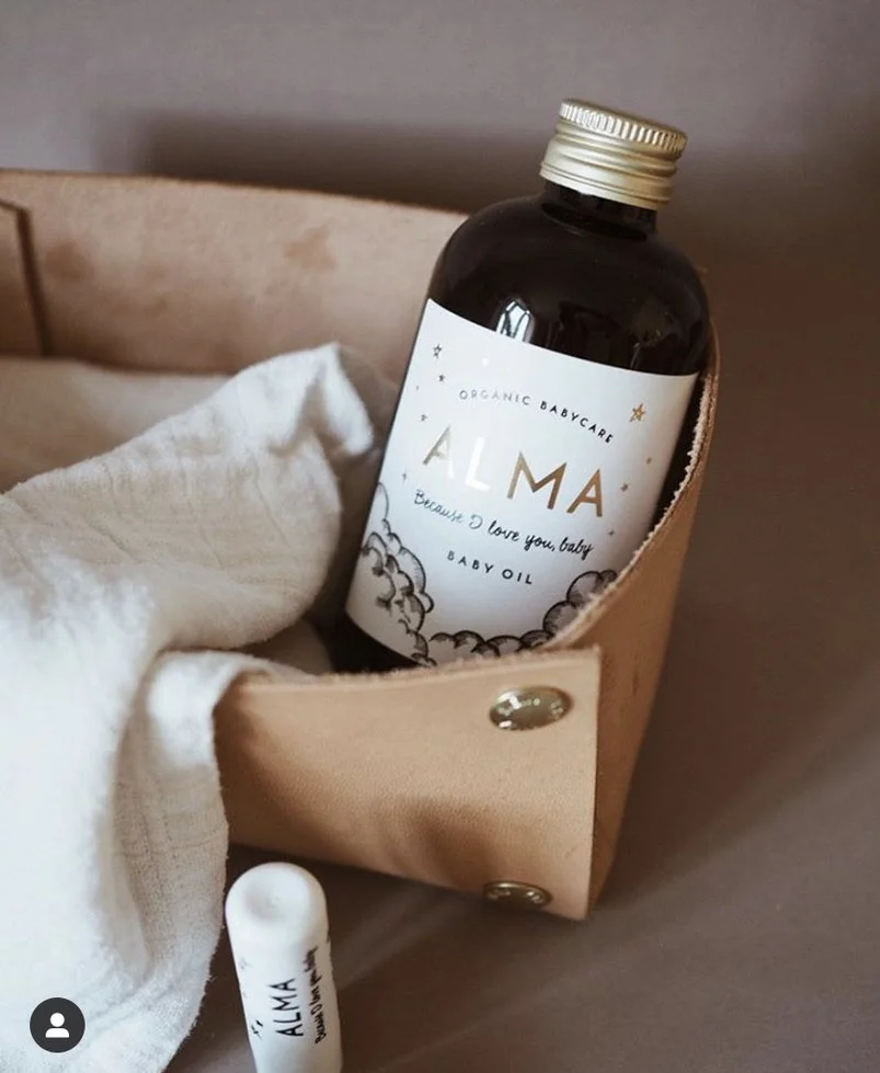 A small bottle of Alma organic baby oil inside a beige leather bag, with a white cloth and a tube of Alma product nearby.