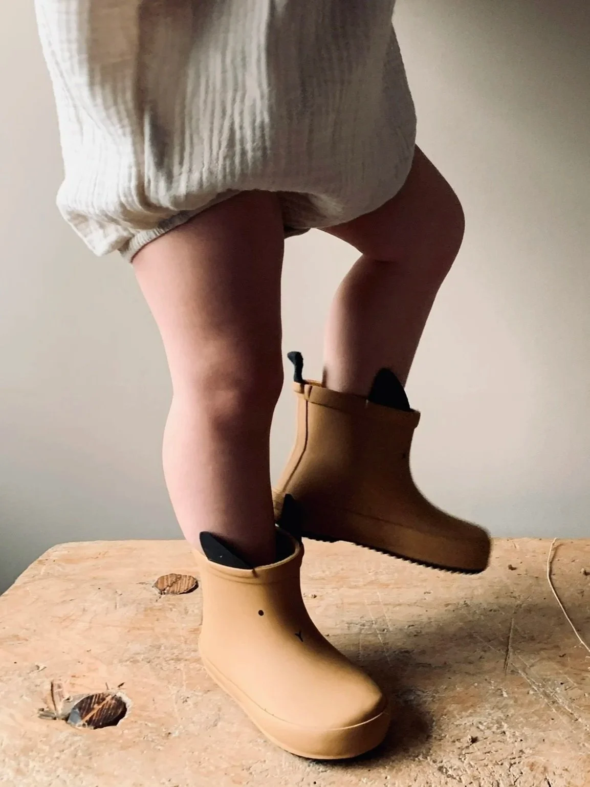 Child wearing beige rain boots, standing on a wooden surface, with one foot slightly off the ground.