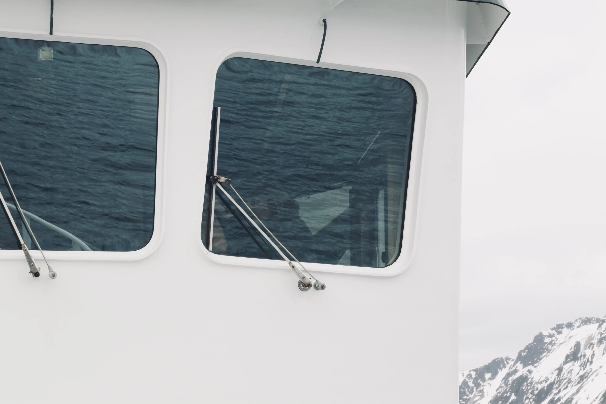 Close-up of a boat's window with a view of the ocean and snow-covered mountains in the background.