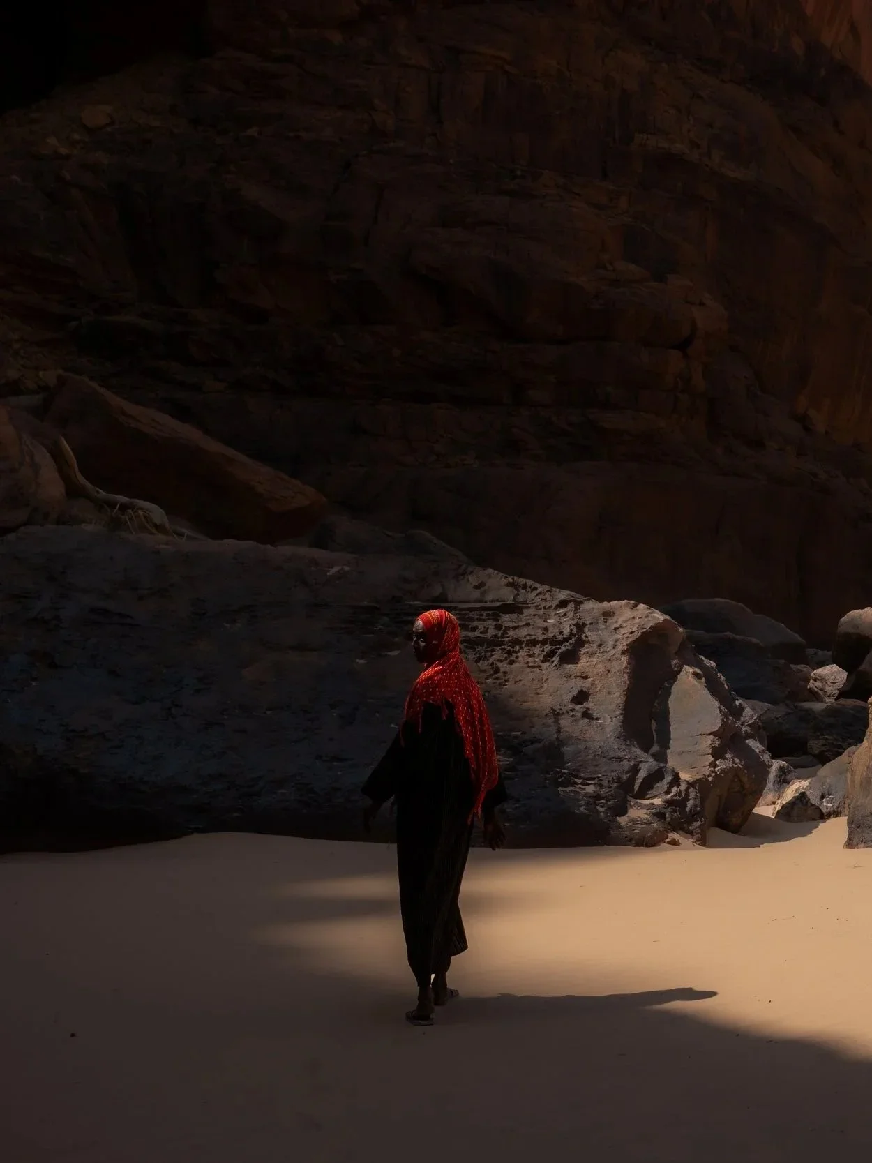 A person dressed in black with a red patterned headscarf walking on sandy terrain inside a deep canyon with high rocky walls.