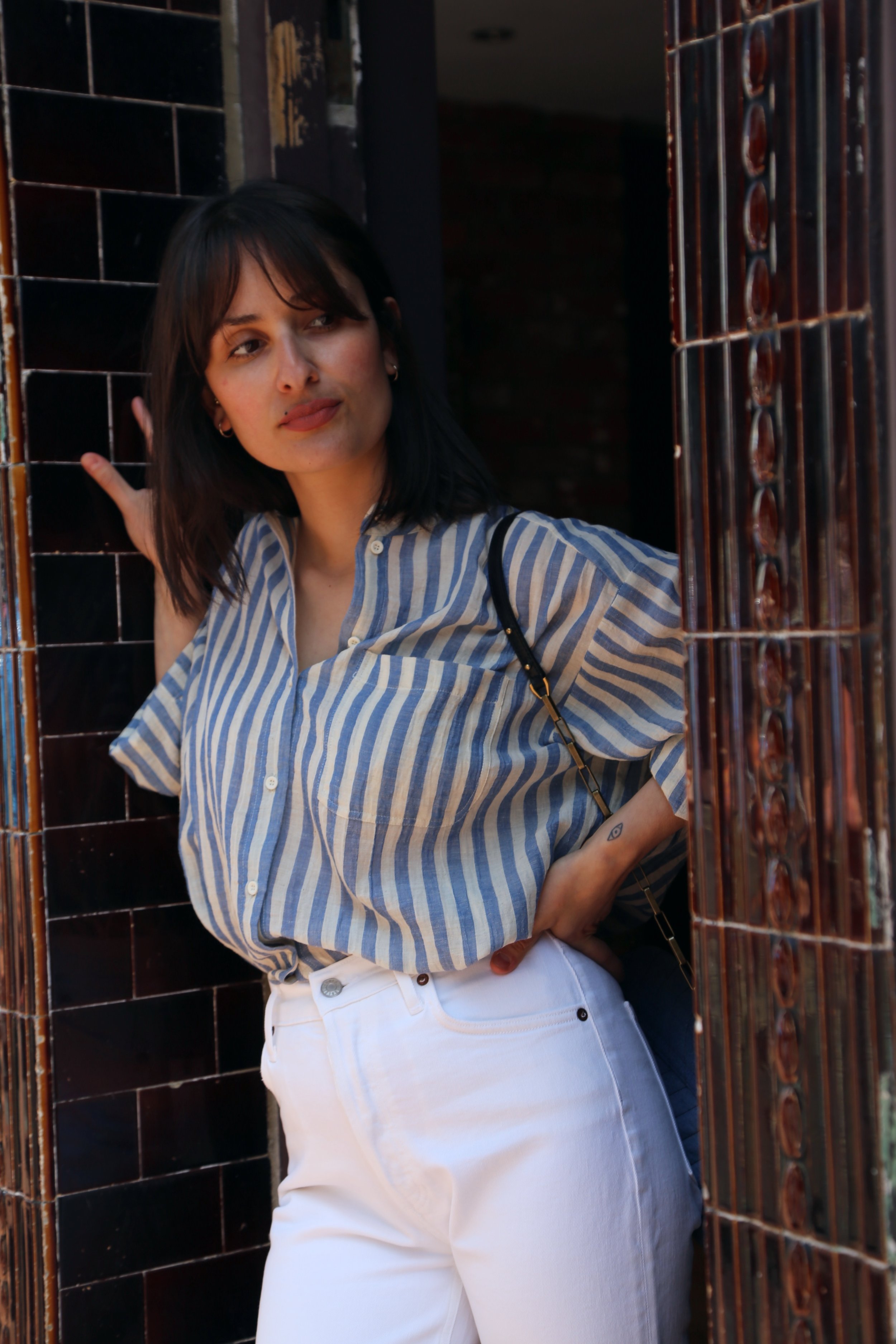 Young woman with dark hair and tattoos, wearing a striped shirt and white pants, standing in a doorway, looking to the side.