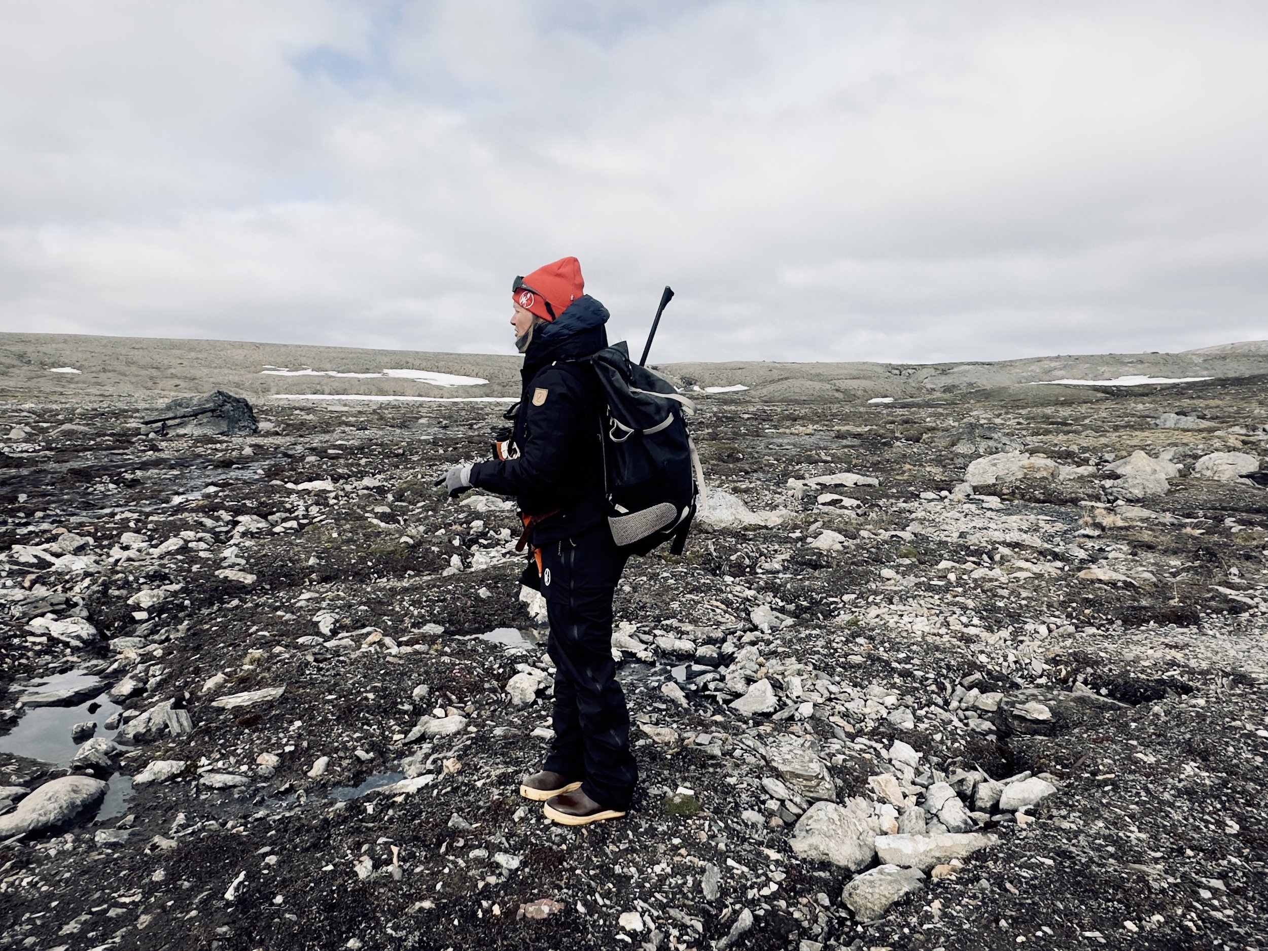 A hiker dressed in black with a red hat and sunglasses standing on rocky terrain in a vast, open landscape with patches of snow and a cloudy sky.