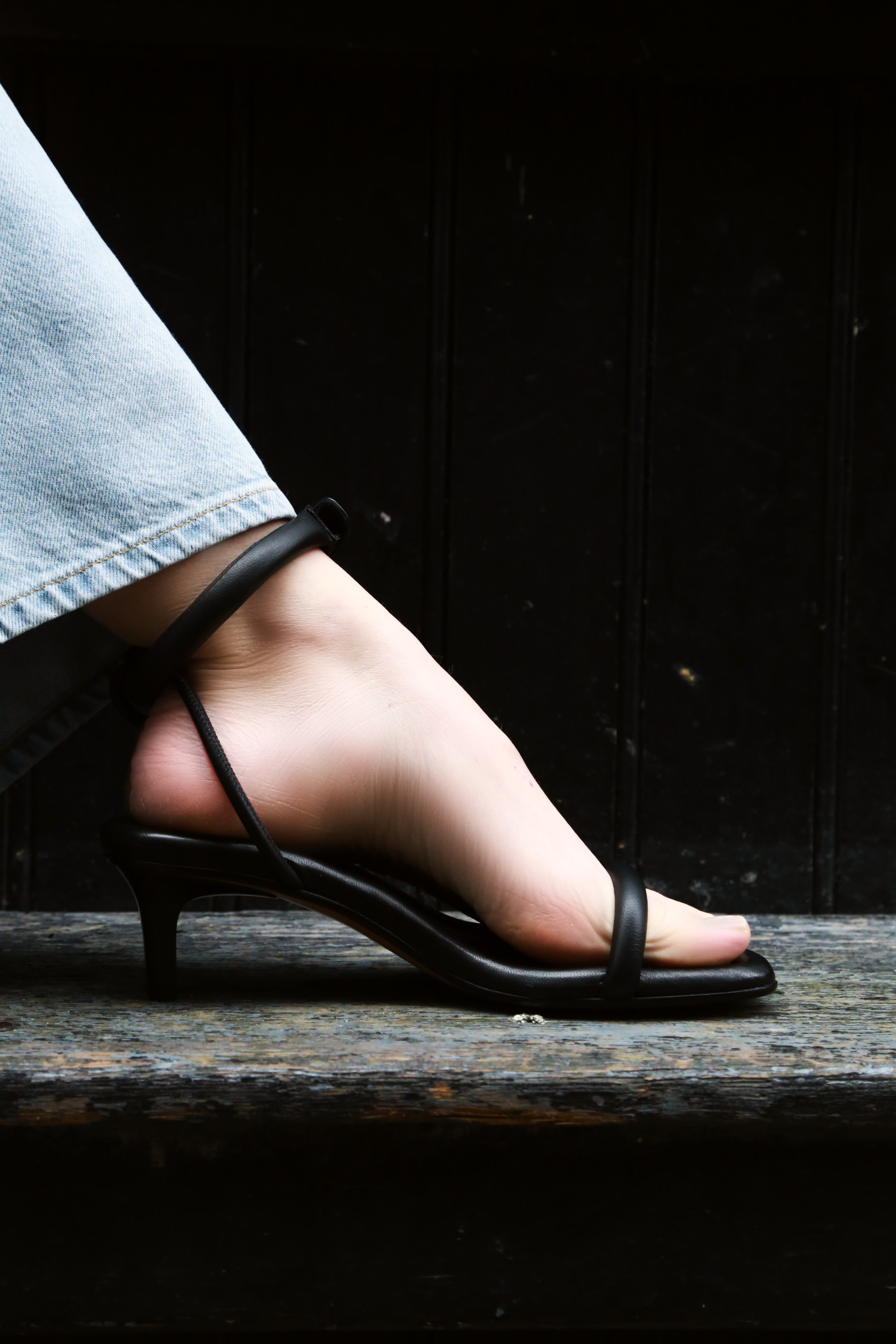 A person's foot in a black high-heeled sandal, resting on a rustic wooden surface with a black background.