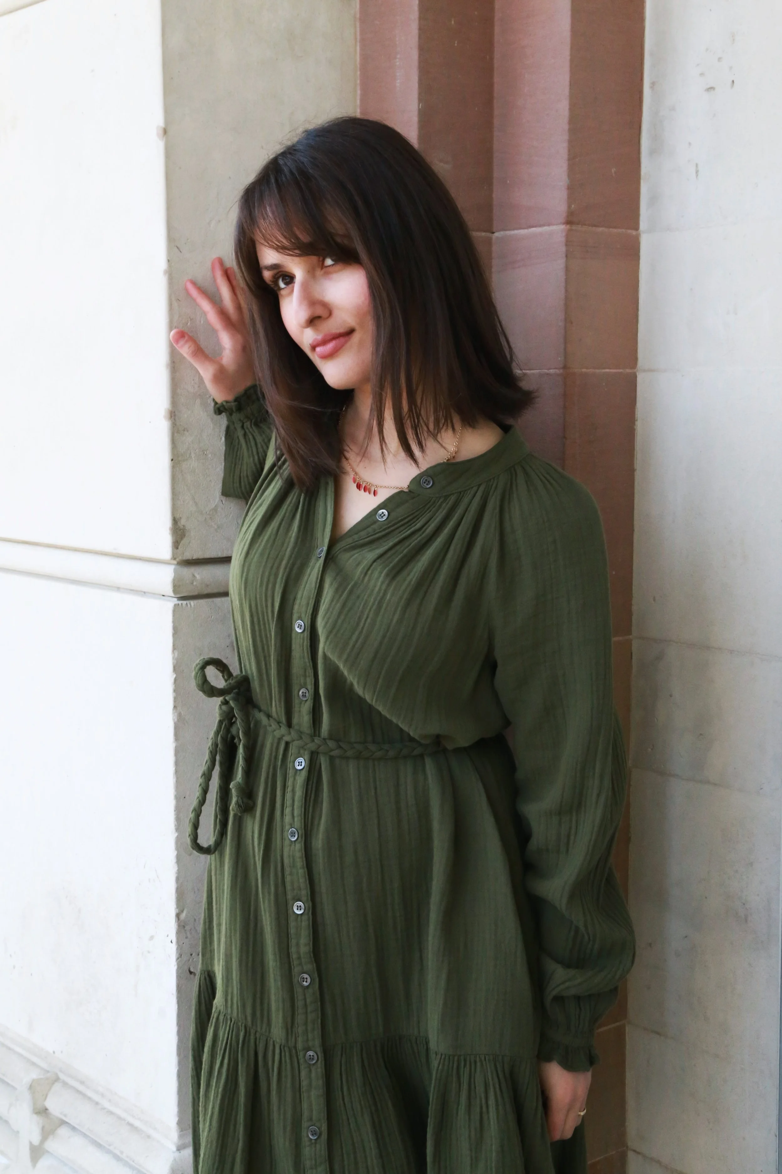 A woman with shoulder-length dark hair in a green dress standing against a beige and brown stone wall, making eye contact with the camera, with a playful expression.