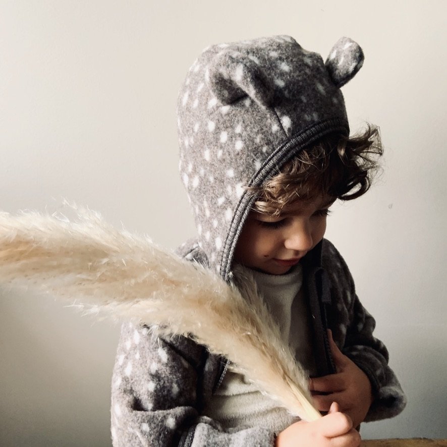 A young child wearing a grey animal-themed hooded jacket with fuzzy ears, holding a large, light-colored feather, standing against a plain white wall.