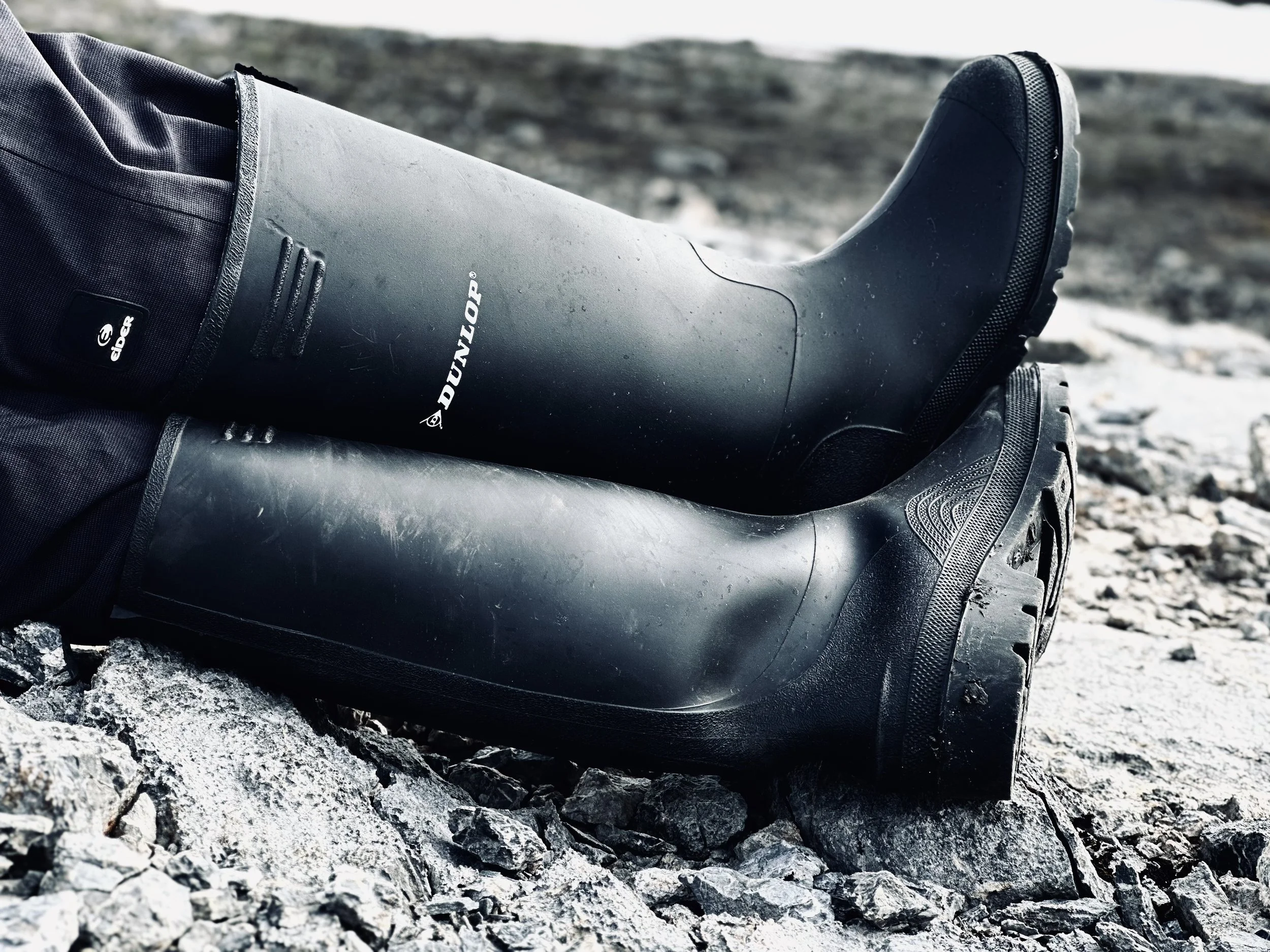 A pair of black rubber rain boots with treaded soles resting on rocky ground outdoors.