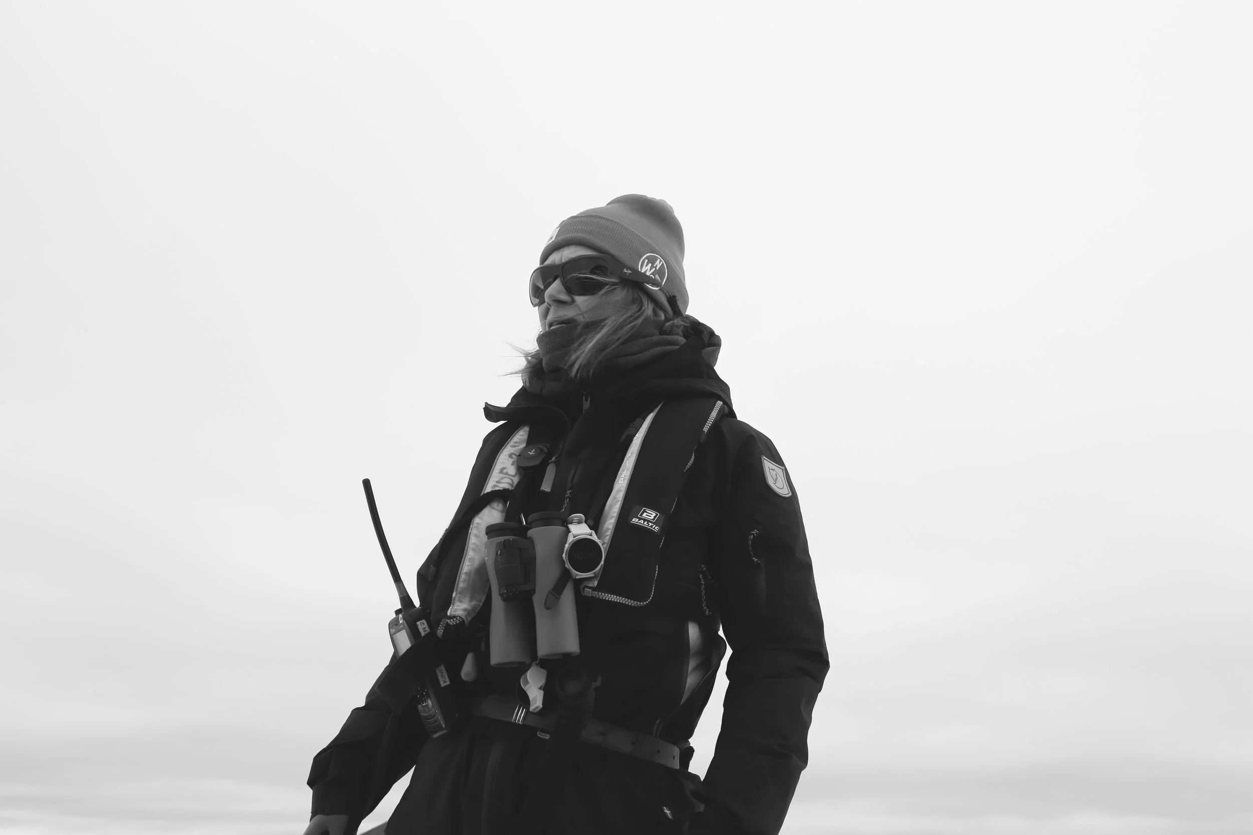 A woman dressed in winter outdoor gear, including a beanie, sunglasses, and a jacket, standing against a gray, overcast sky.
