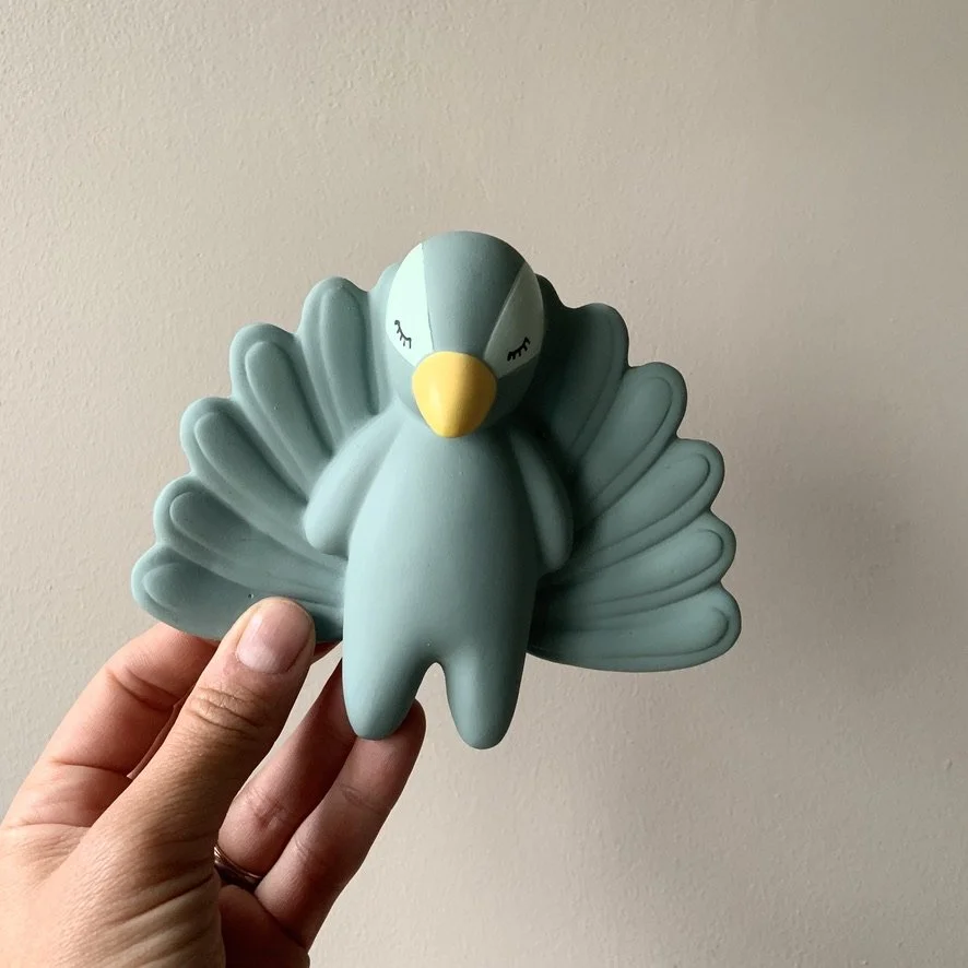 A hand holding a blue ceramic bird-shaped figurine with a yellow beak and closed eyes, positioned against a plain beige wall.