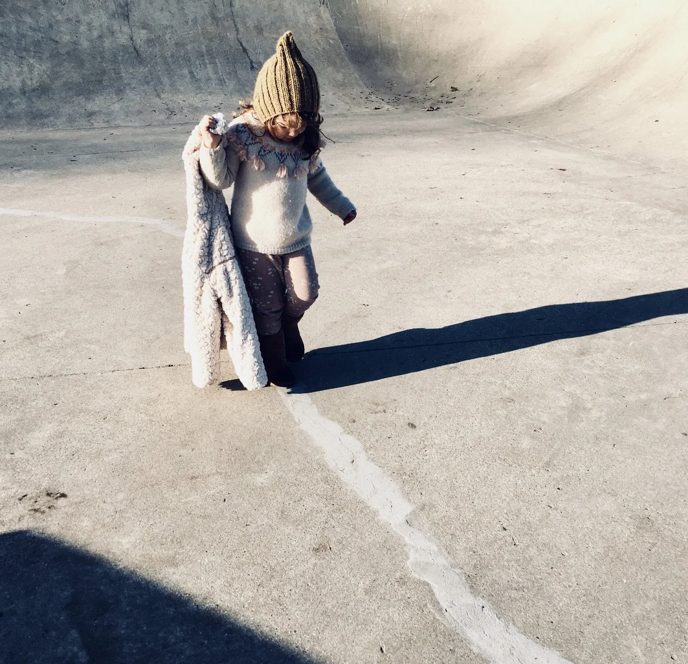 A young girl wearing a tan knit hat, a beige sweater with playful design, pink pants, and black boots, is standing on concrete in a skatepark and drawing a line on the ground with her finger, casting a long shadow.