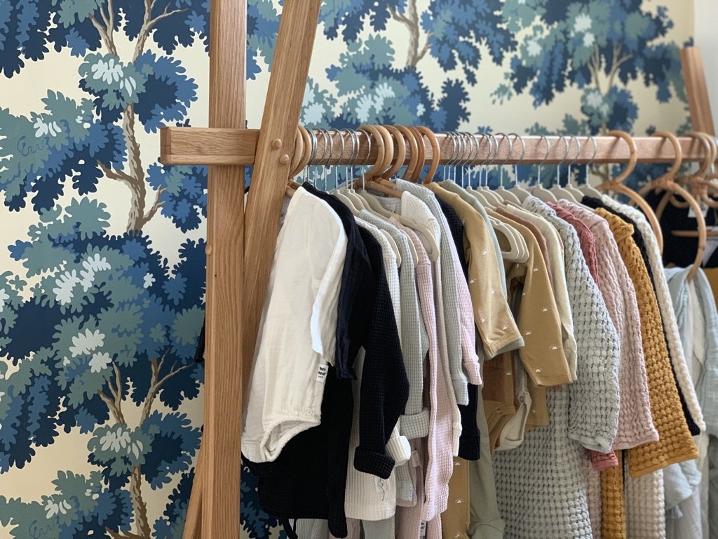 A wooden clothing rack holding various baby or toddler clothes, with a blue and white leafy patterned wall in the background.