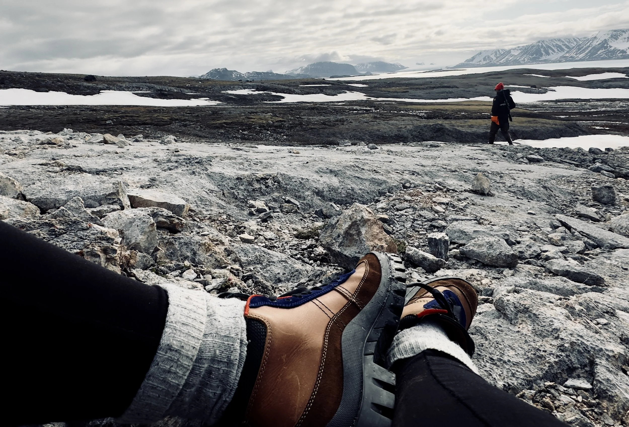 View of a rocky, barren landscape in a cold, mountainous area with patches of snow, showing a person in hiking boots and socks resting with legs extended, and someone walking in the distance.