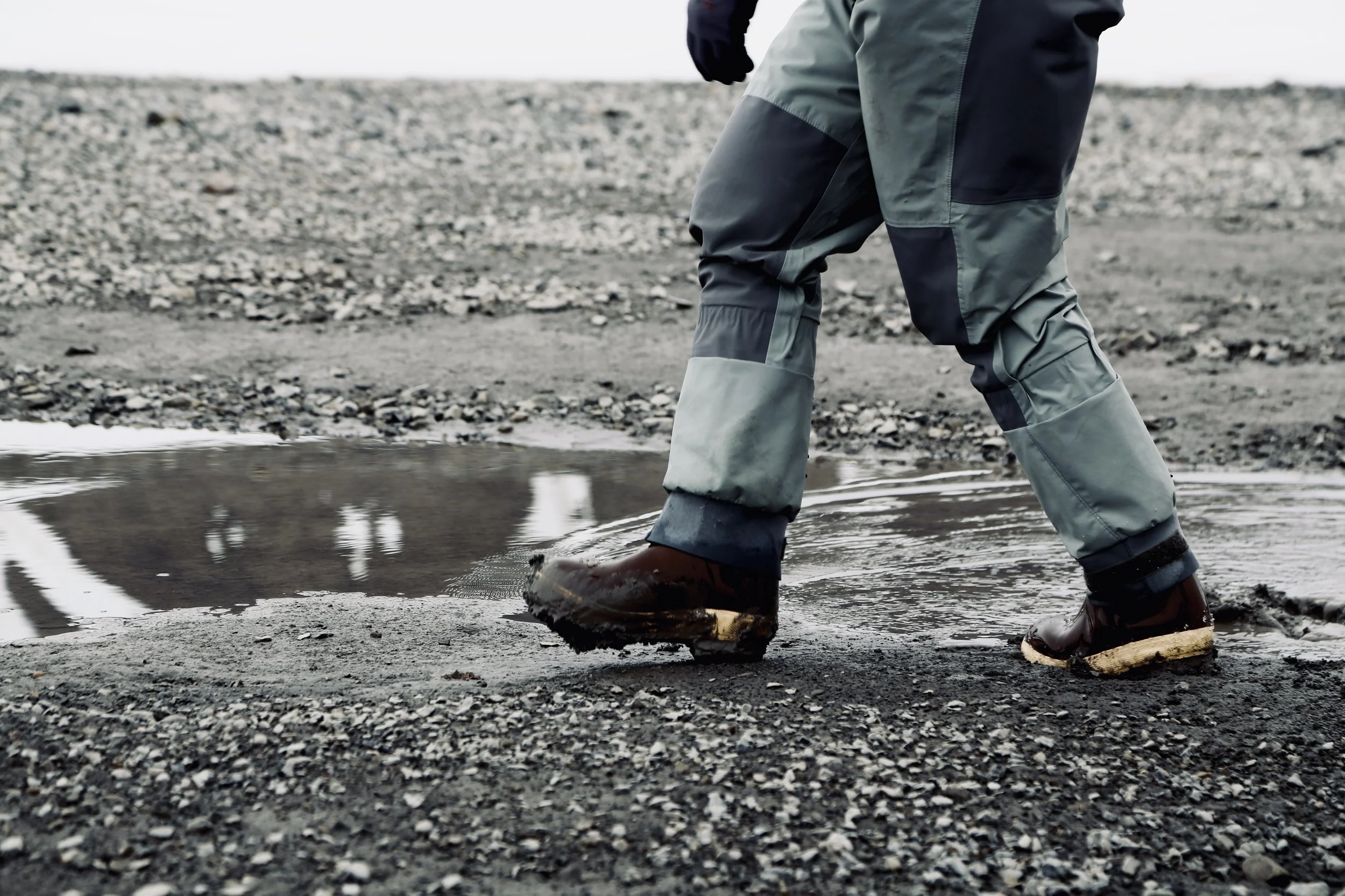 Person wearing waterproof boots and outdoor gear stepping into a small puddle on a gravelly surface.