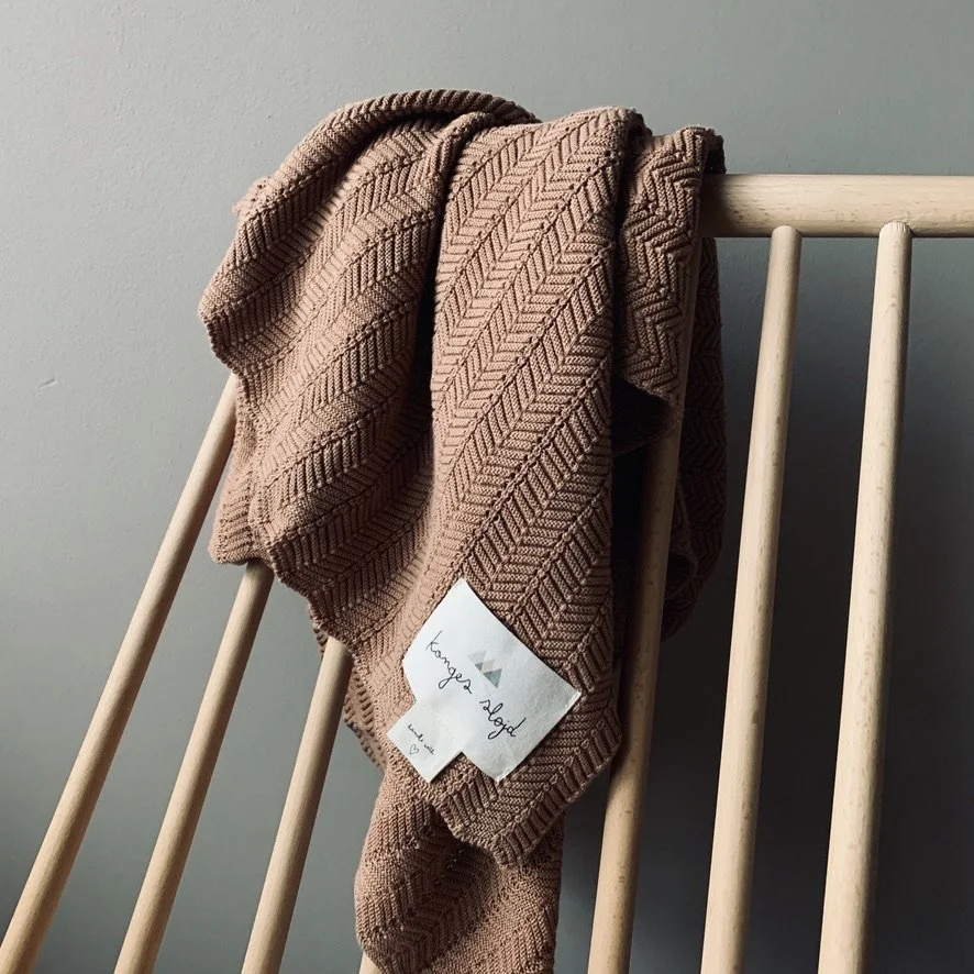 A beige textured blanket or scarf draped over a wooden crib or bed rail, with a white clothing tag attached.