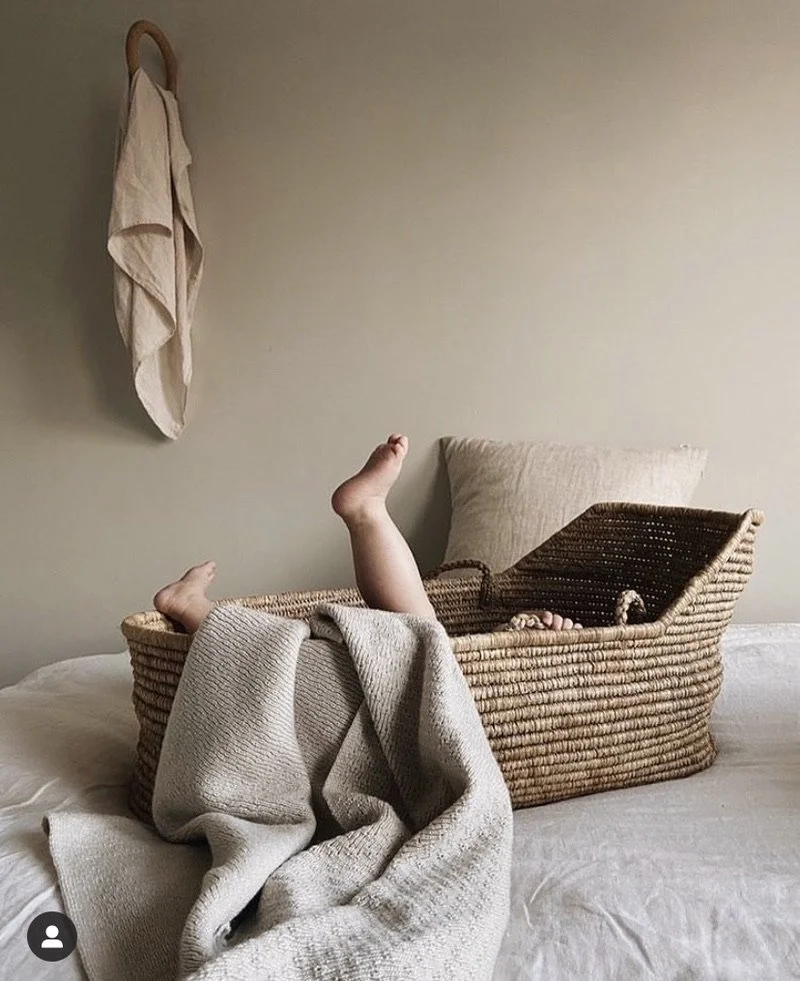 A wicker basket with a child inside, showing only their legs and feet, on a bed with beige and cream-colored bedding. A beige blanket drapes from the basket's side.