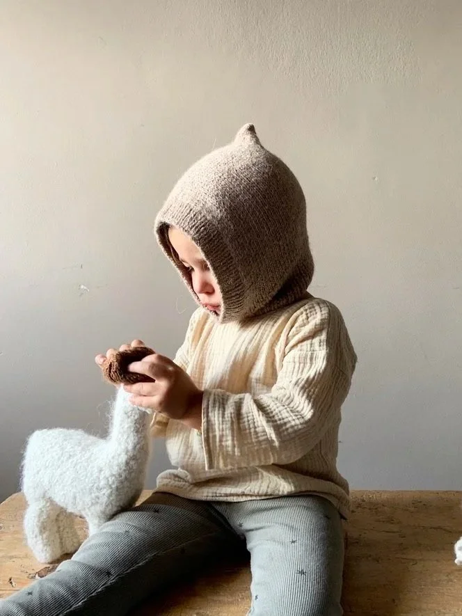 Child wearing a beige hoodie with a pointed top, sitting on a wooden surface and holding a small stuffed animal, with a neutral plain wall in the background.