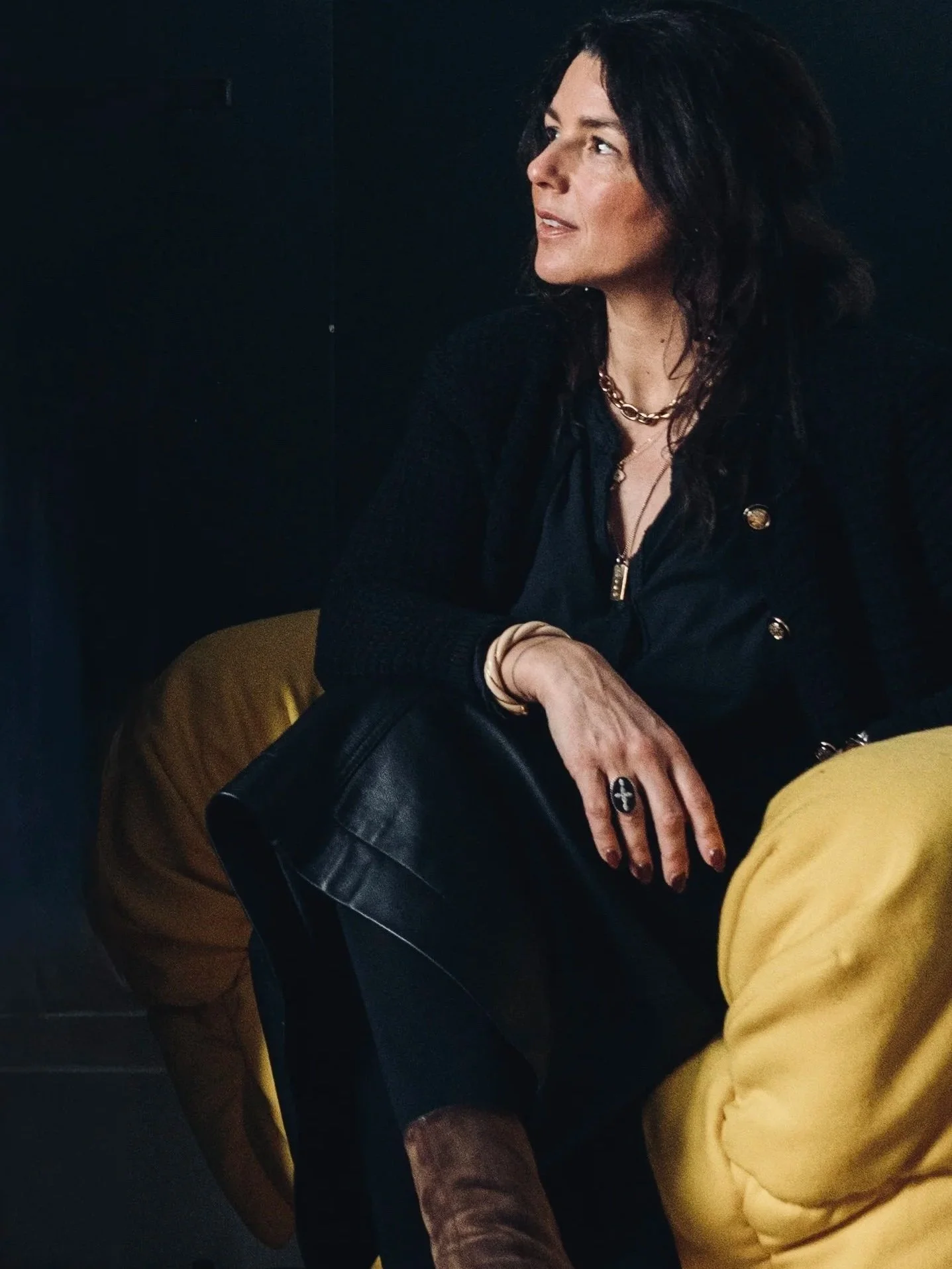 A woman with dark hair wearing a black shirt, gold jewelry, and yellow pants, sitting in a dark room with a black background.