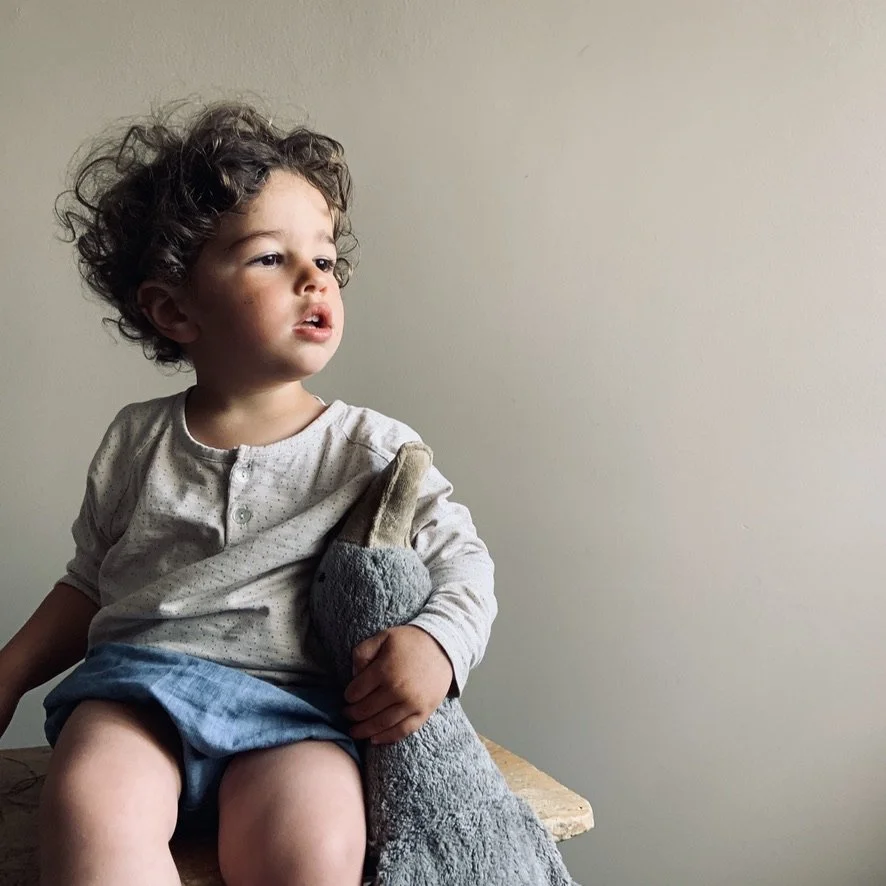 A young child with curly hair sitting on a bench, holding a stuffed rabbit toy, looking off to the side against a plain wall.