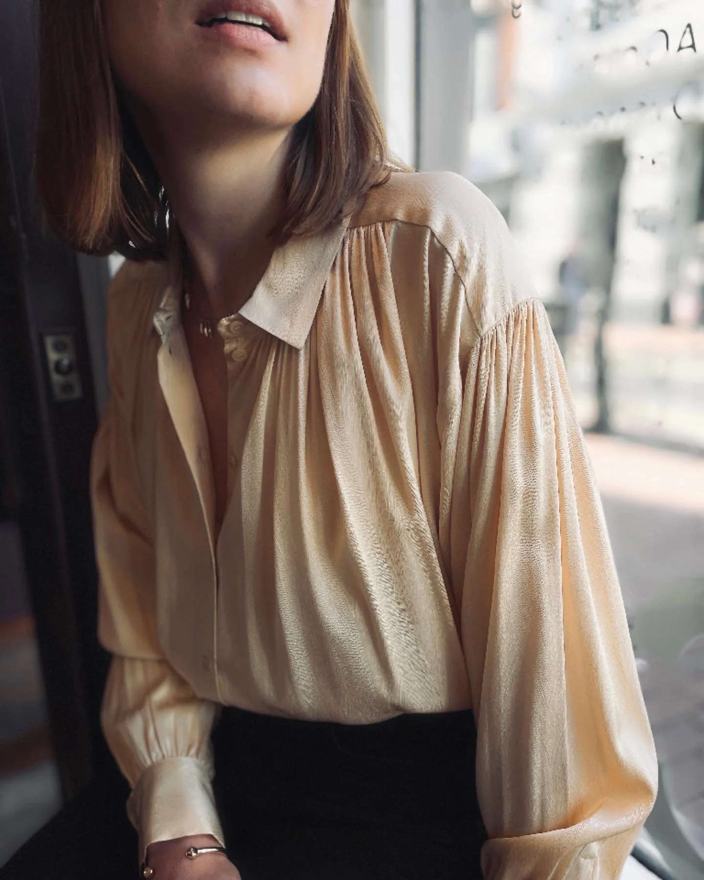 Close-up of a woman wearing a shiny cream-colored blouse with pleated sleeves, seated near a window with sunlight, with her face partially visible.