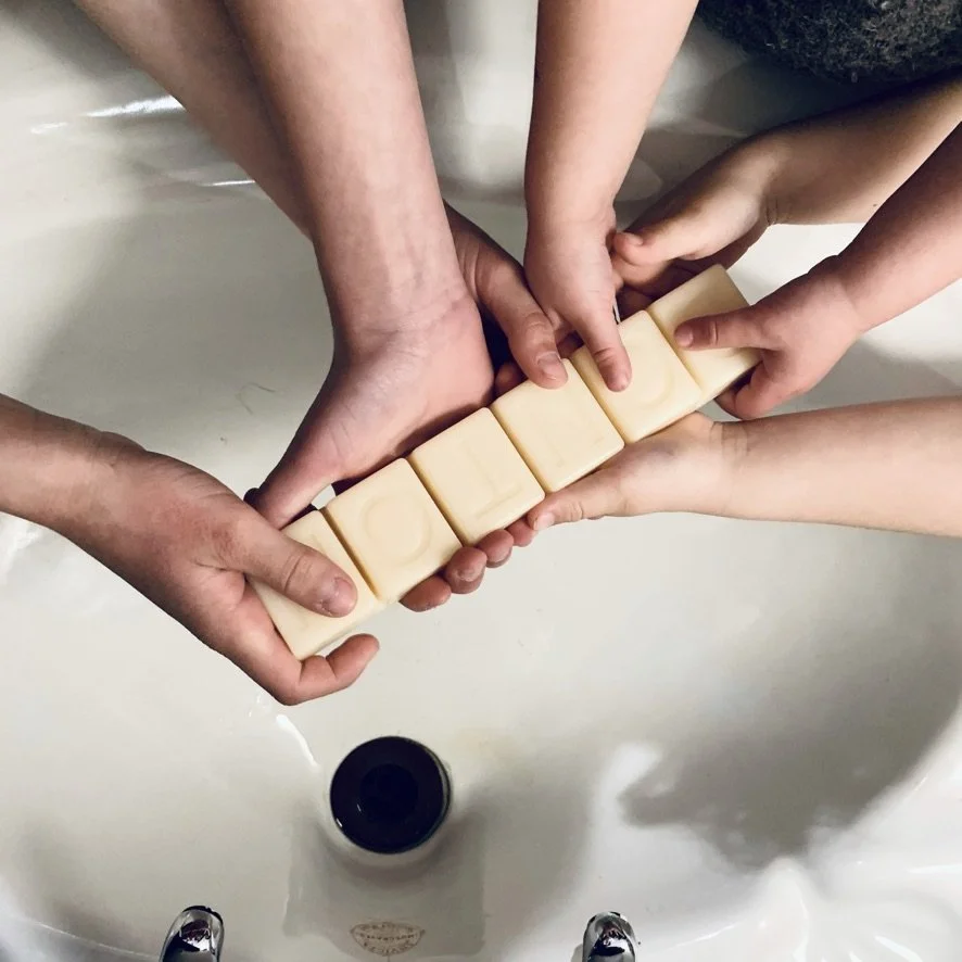 Multiple hands holding bars of soap spelling out 'LAUNDRY' over a sink.