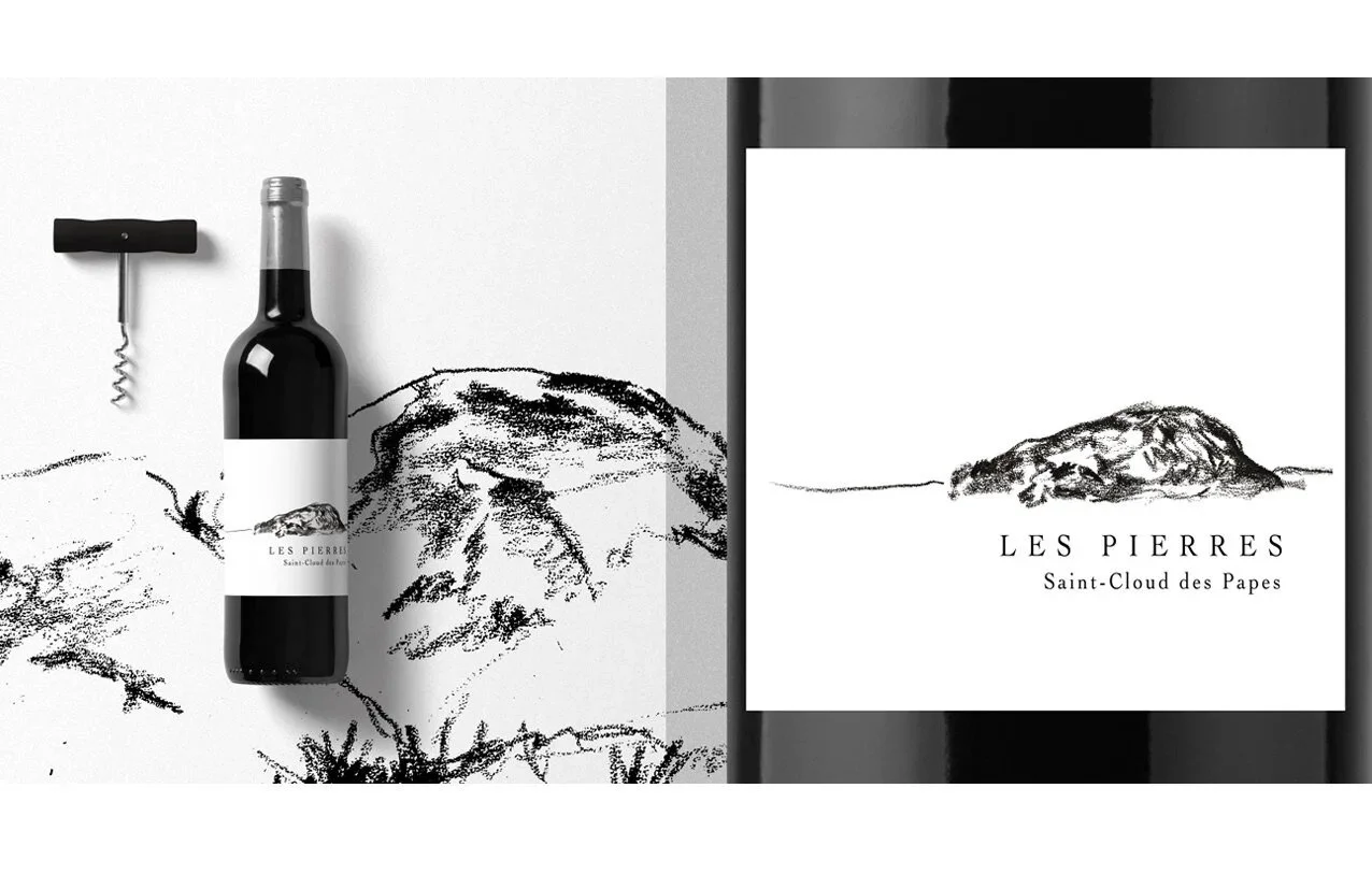 A wine bottle with a label that reads 'Les Pierres Saint-Cloud des Papes' positioned on a white surface next to a corkscrew. The surface is decorated with a black ink sketch of mountainous terrain and rocks, matching the illustration on the wine bott