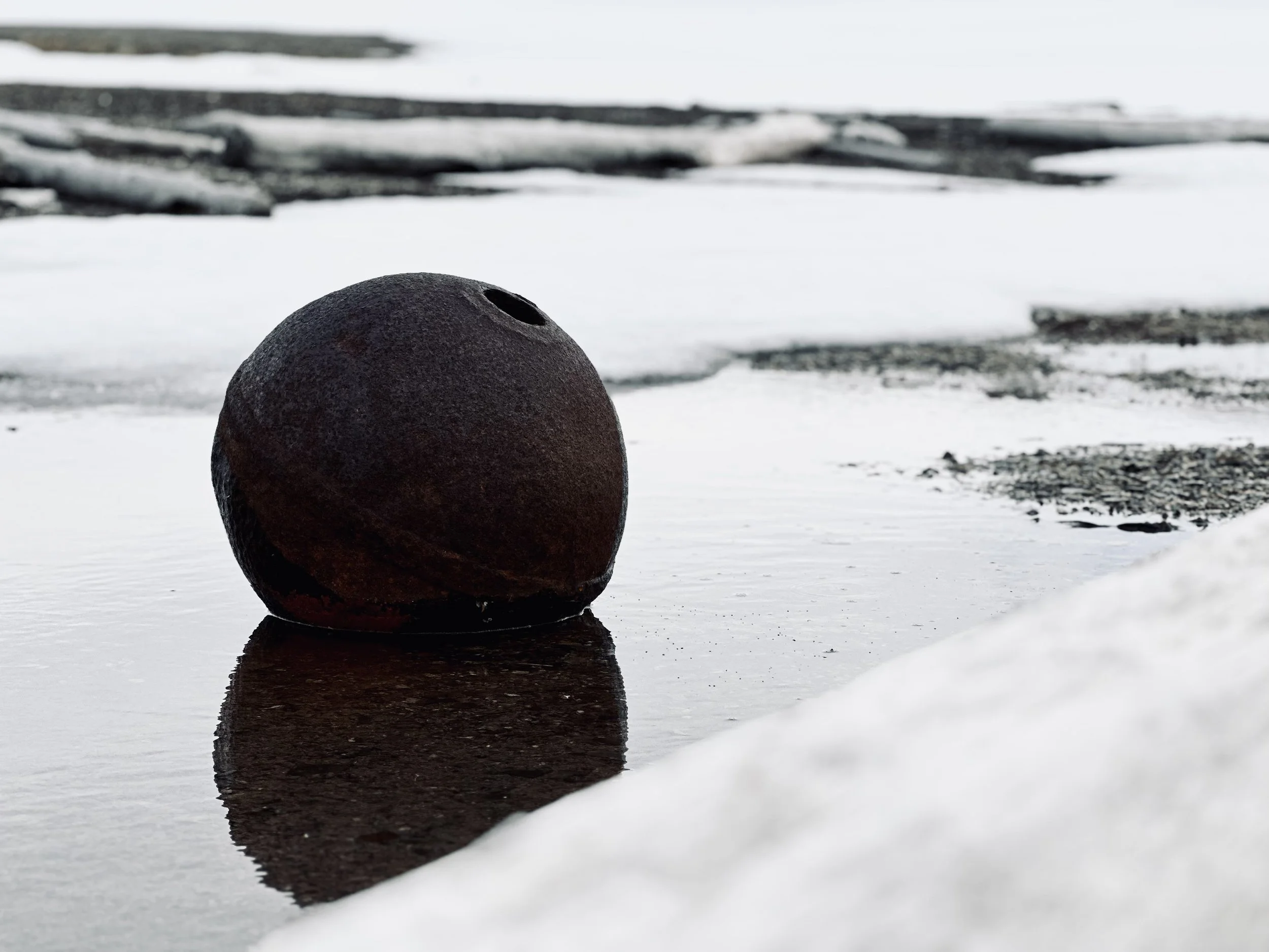 A rusted, old, spherical bomb rests on a reflective icy surface with snow and ice around it and in the background, overcast sky.