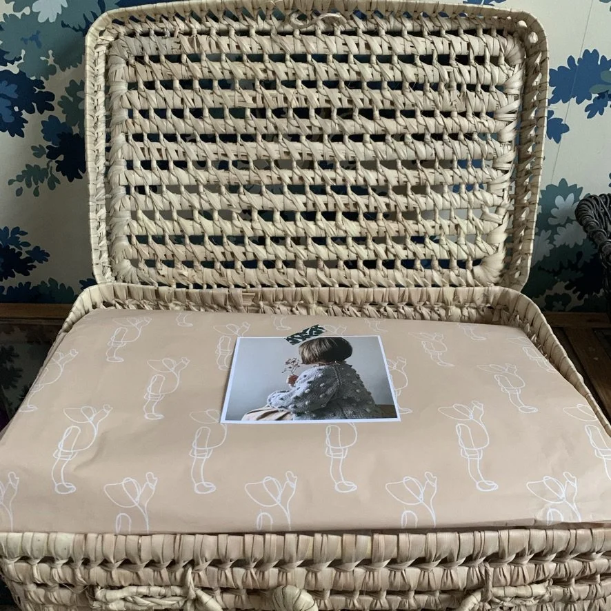 A wicker basket with a lid open, lined with patterned paper that has white line drawings of a child with a balloon. A printed photograph of a child with short hair, wearing a patterned top, is placed on the paper inside the basket.