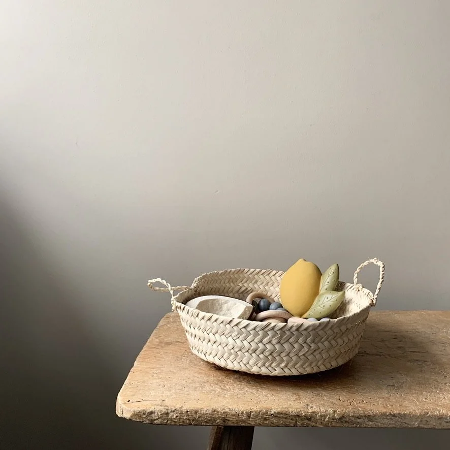 A woven basket on a wooden table containing various decorative ceramic fruits including a banana, an apple, and a lemon along with some small rocks.