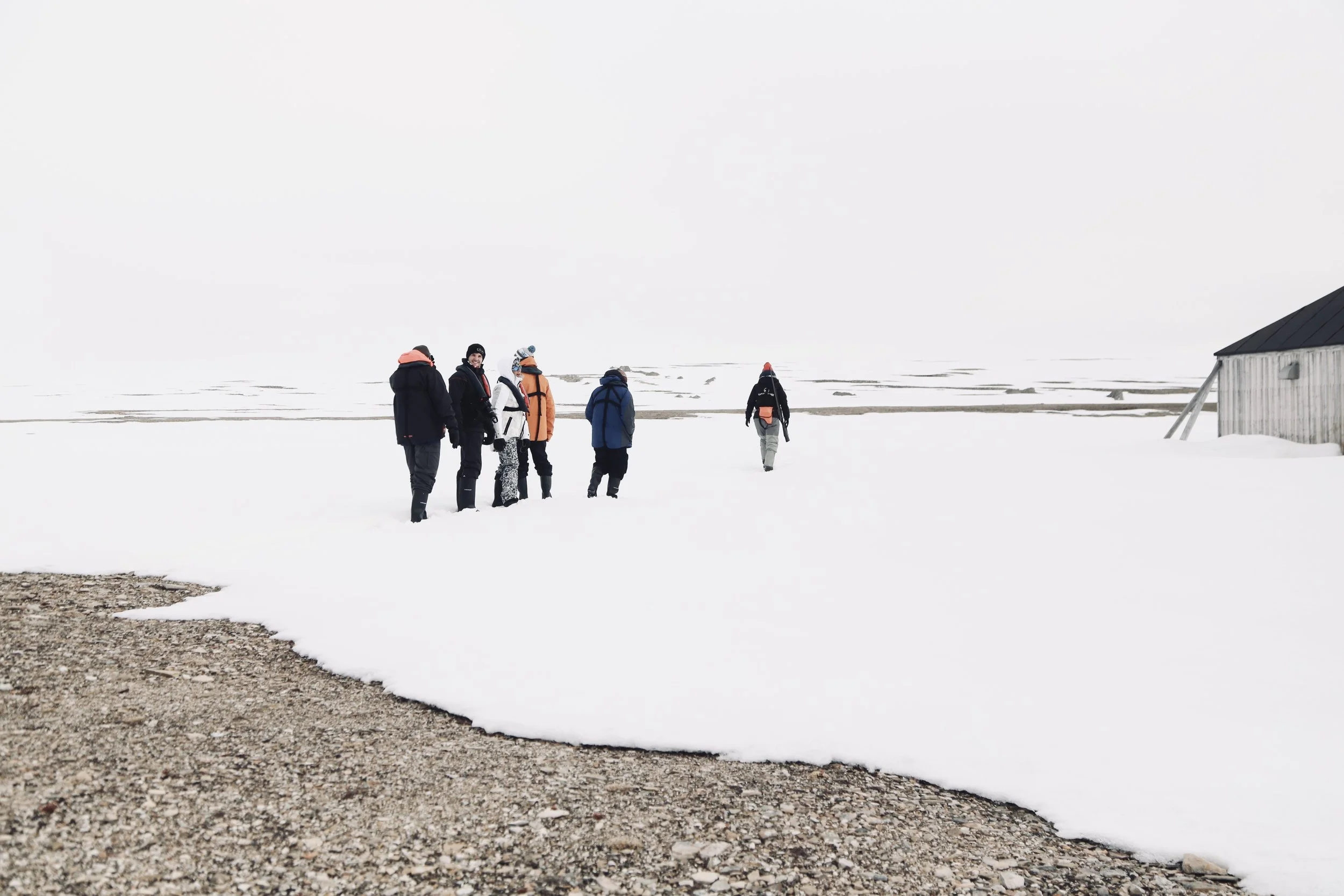 Group of six people hiking on snow-covered landscape with a small building on the right.
