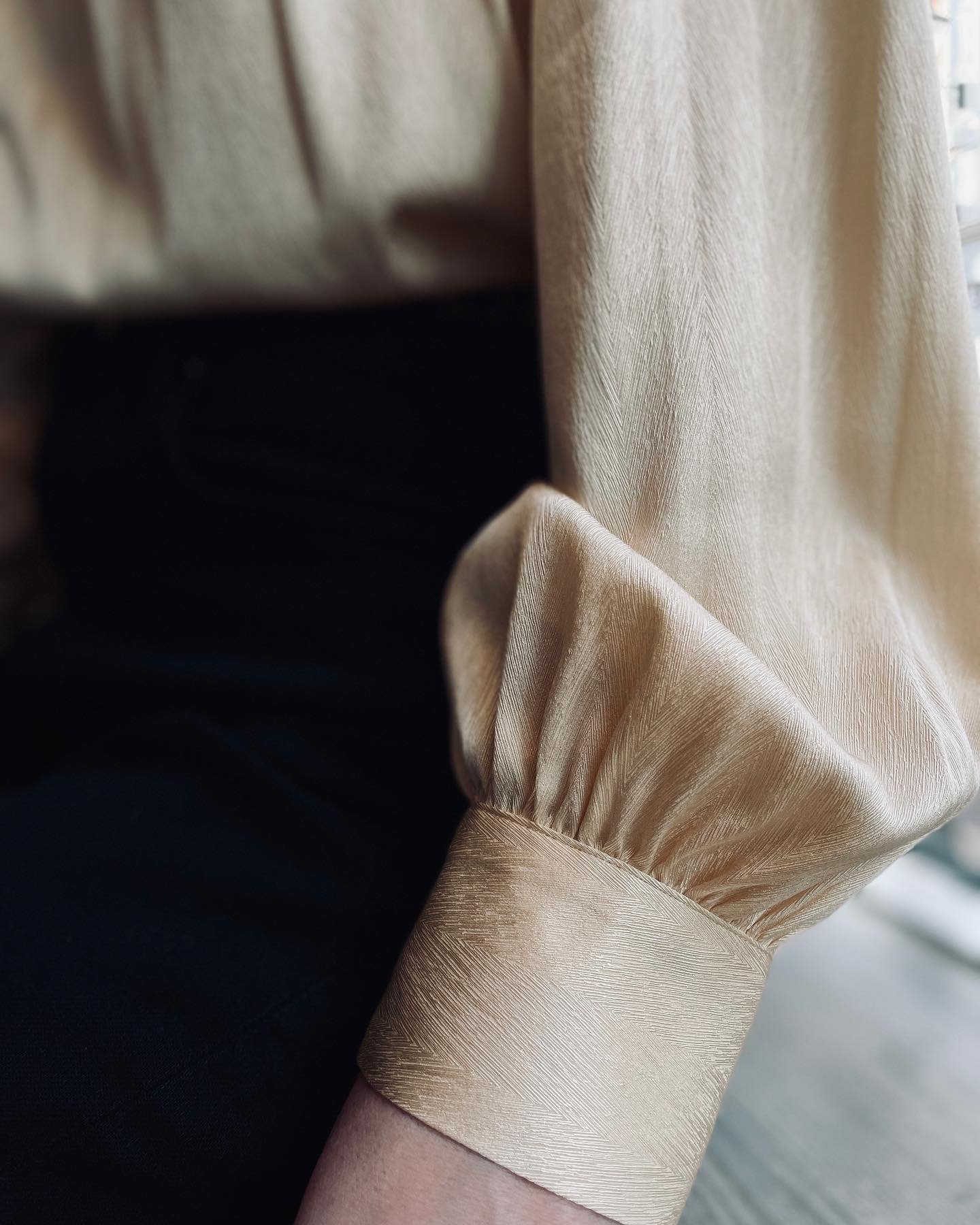 Close-up of a person wearing a beige silk blouse with puffed short sleeves and black pants, next to a window with beige curtains.