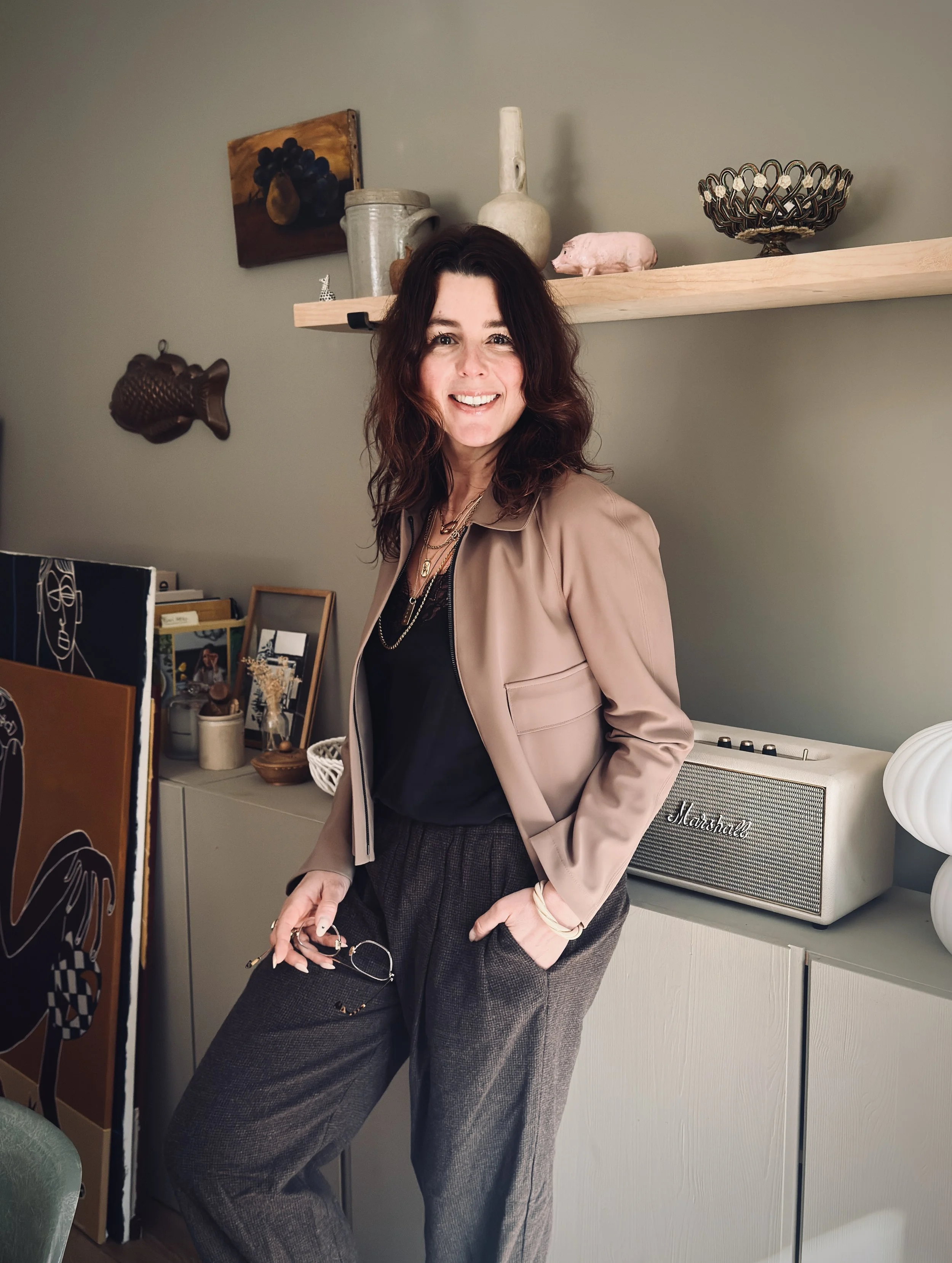 A woman with shoulder-length wavy brown hair smiling in a modern living room, wearing a beige blazer over a black top and gray tailored pants, holding glasses in her left hand, standing next to a light-colored cabinet with decorative items and a Marshall speaker.