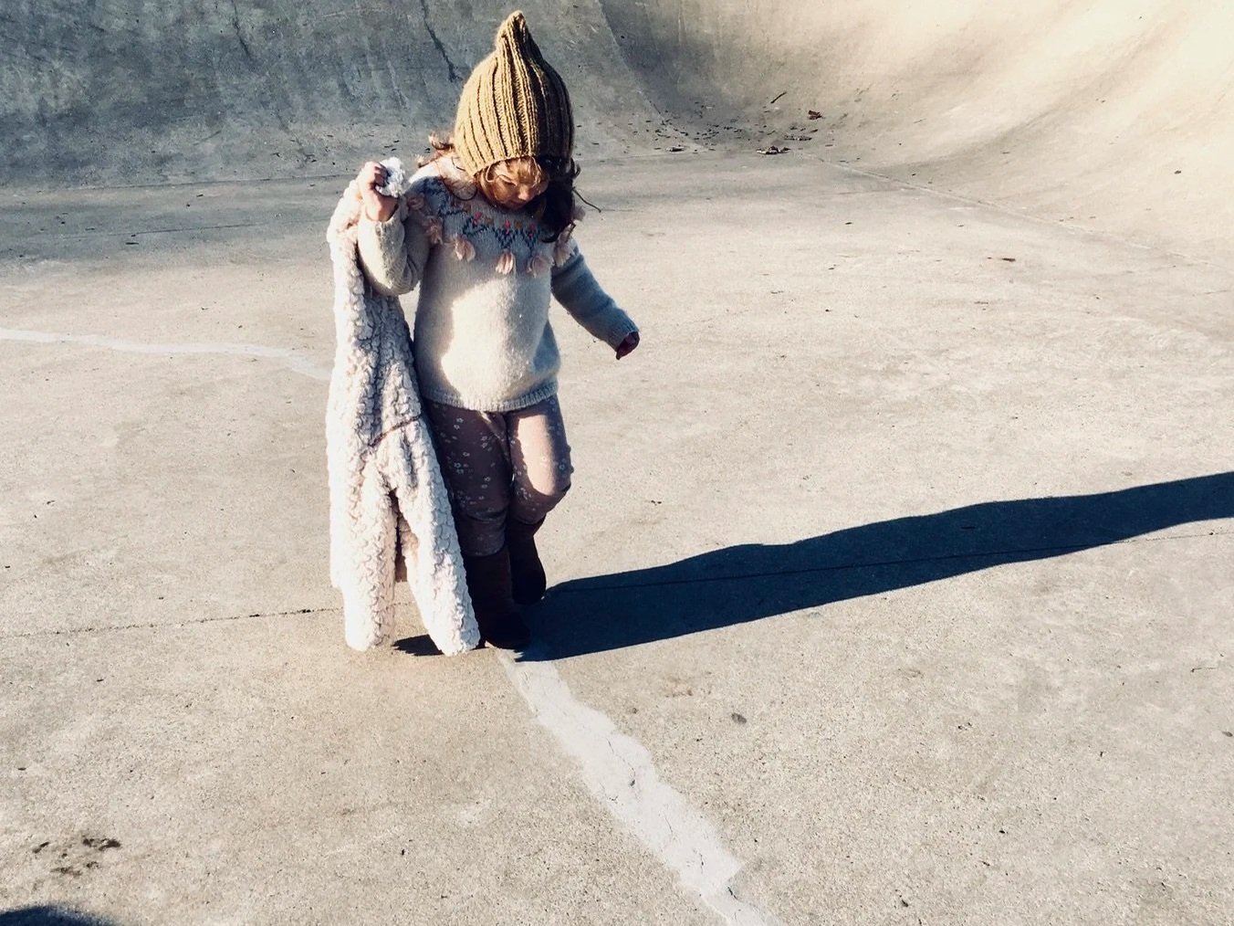 Child wearing a warm sweater, pants, and a beanie hat, walking alone on a large, empty concrete surface, casting a long shadow in the sunlight.