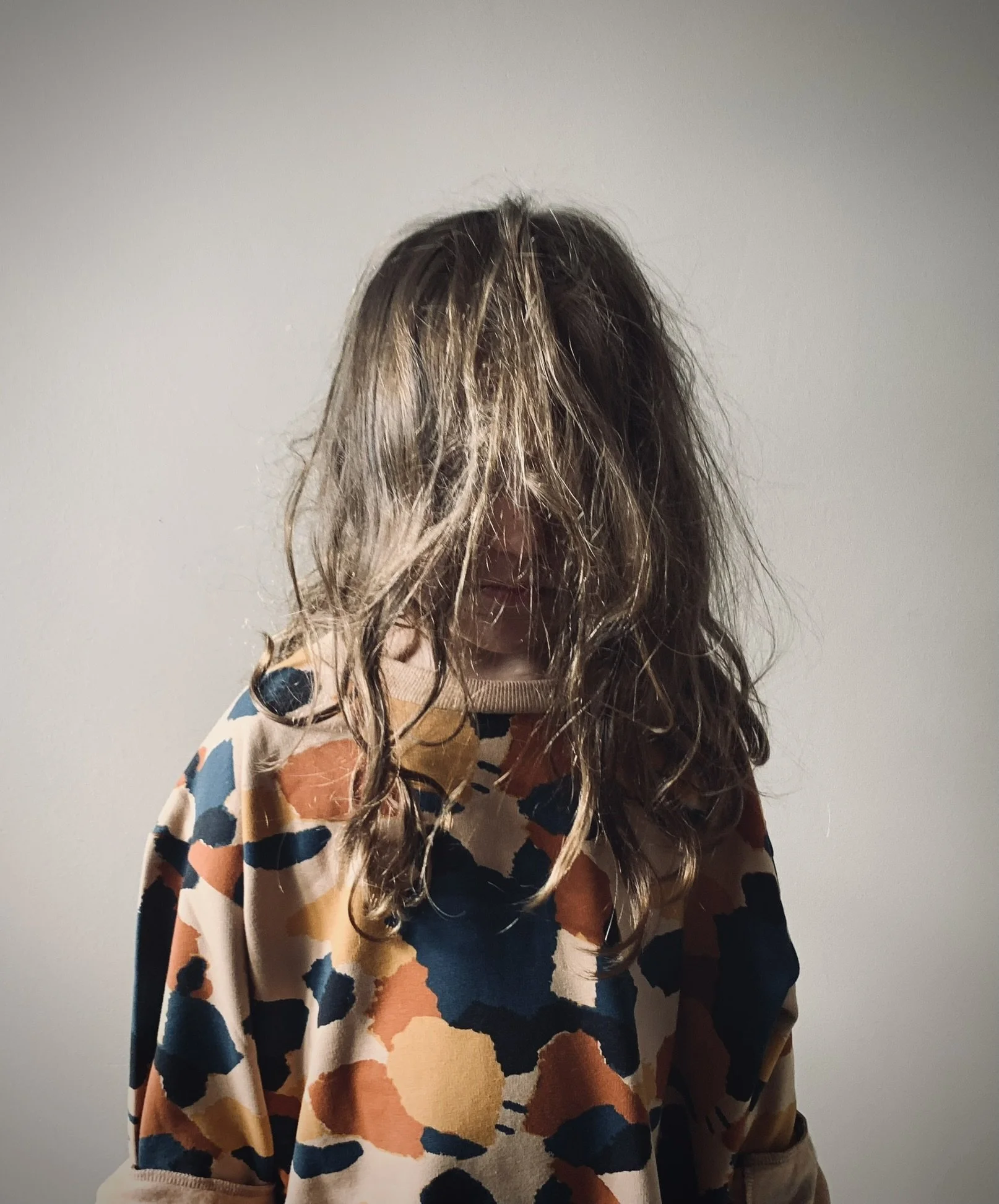 Person with long, wavy, light brown hair covering face, wearing a multicolored patterned sweater, standing against a plain white wall.