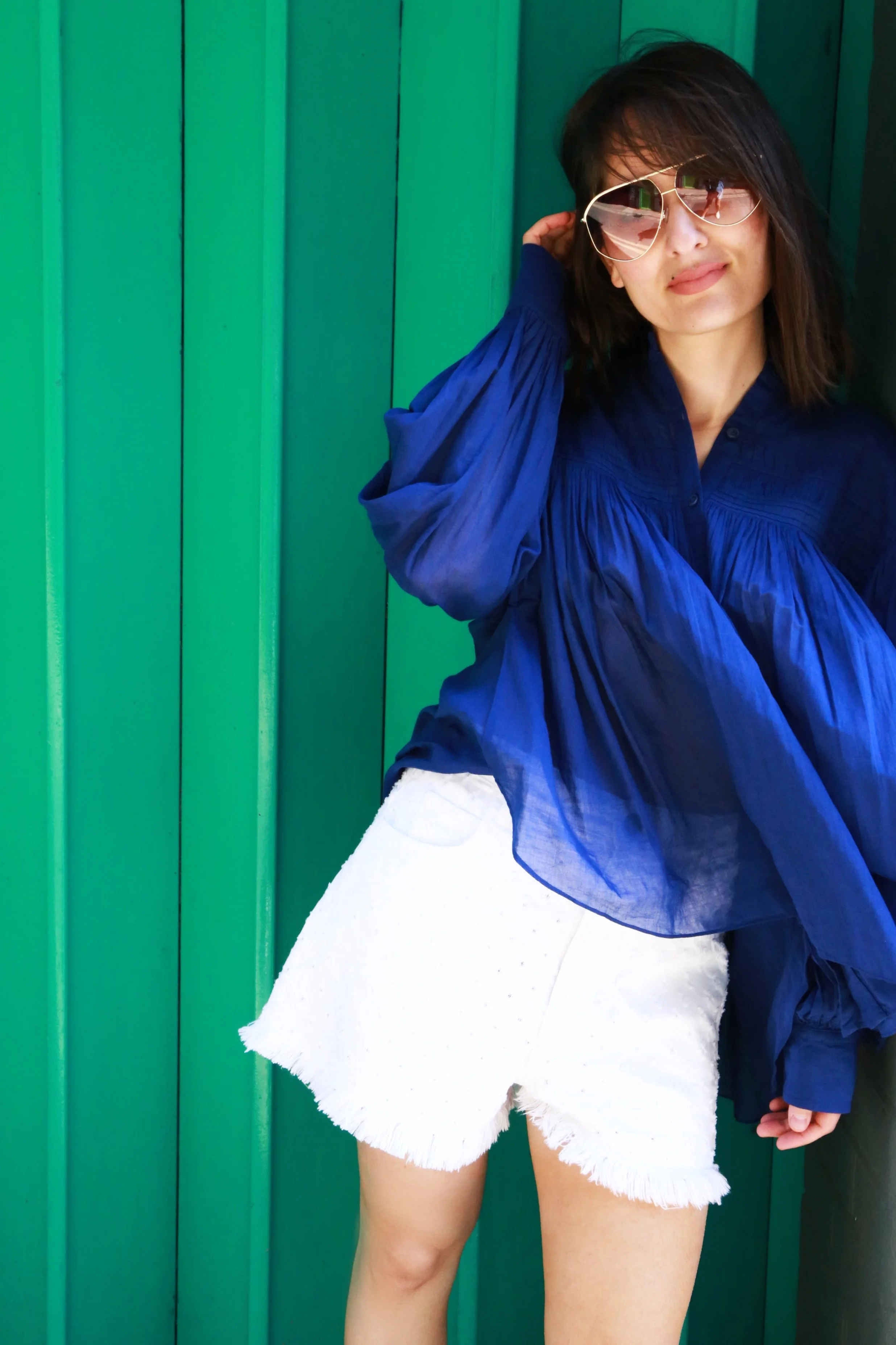 A woman with dark hair wearing sunglasses, a blue flowing blouse, and white denim shorts, standing against a green wooden wall.