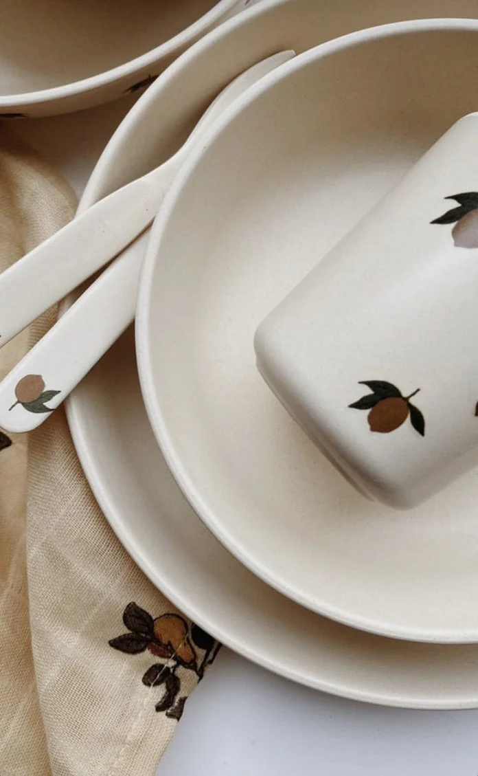 Set of cream-colored ceramic tableware with a lemon and leaf design, including plates, bowls, a butter dish, and spoons, arranged on a linen cloth.