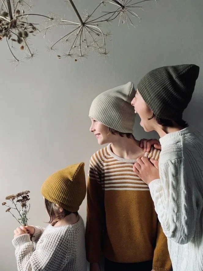 Three children wearing knit hats, standing together indoors, with the eldest smiling at the middle child, and the youngest holding a bouquet of dried flowers.