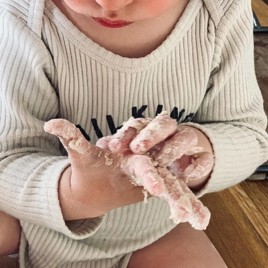 A child in a beige sweater with black lettering has hands covered in flour, holding dough or bread dough.