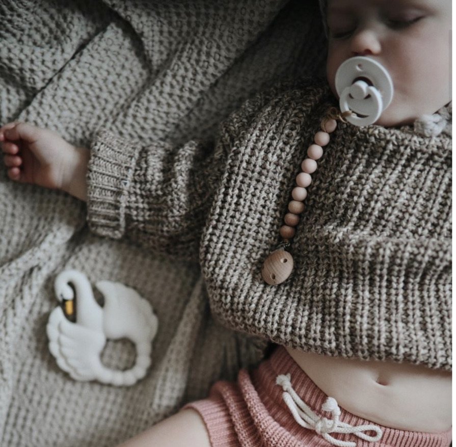 Close-up of a sleeping baby wearing a brown knitted sweater with a pacifier in their mouth. The baby has a beaded necklace and visible clothes. The baby is lying on a textured beige blanket.