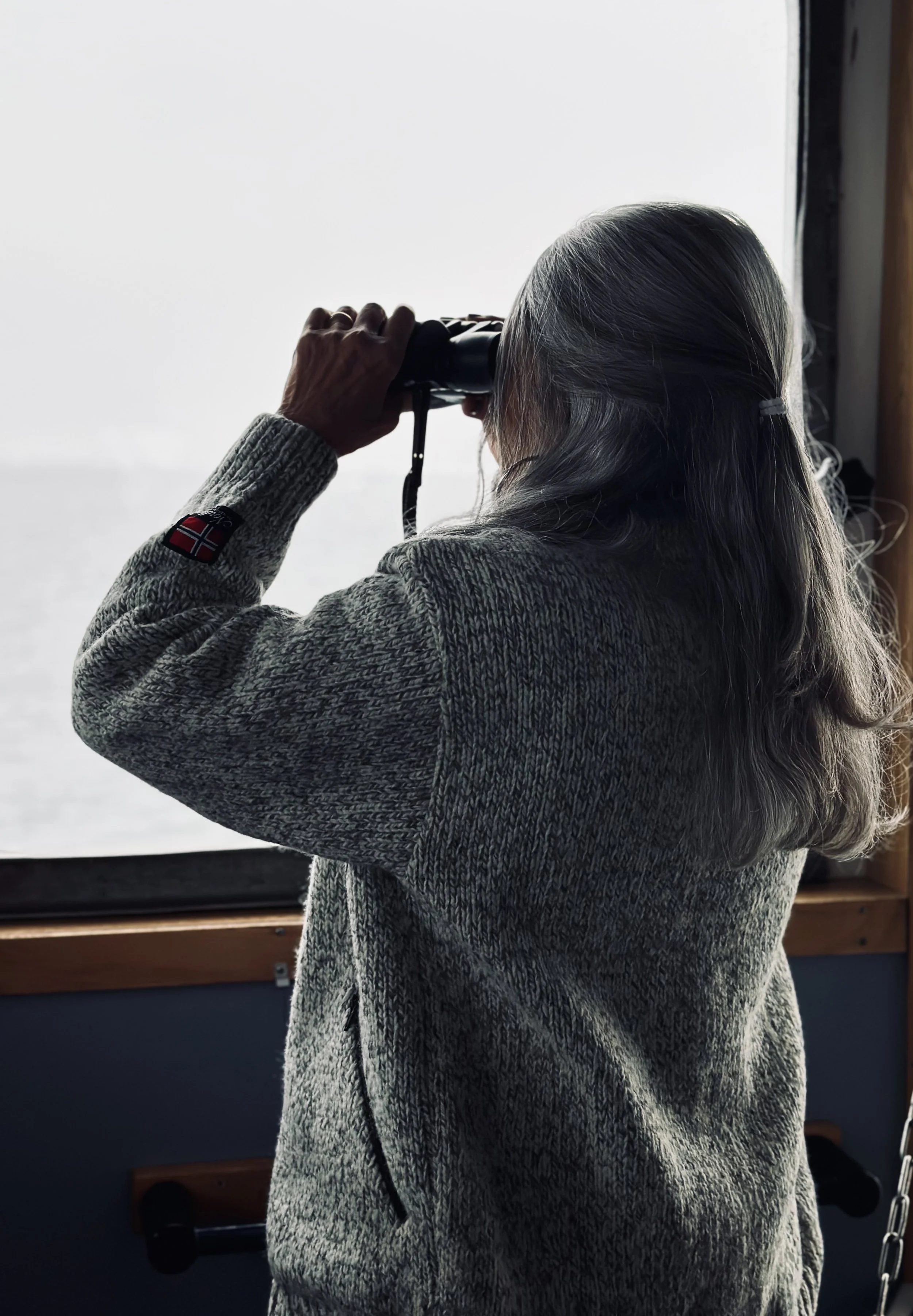 Older woman with gray hair looking through binoculars out a window.
