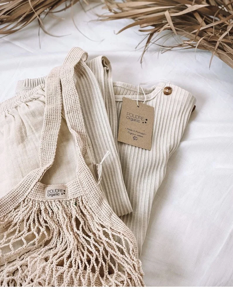 Clothing items made of organic fabrics, including a fringed knit bag, a ribbed long-sleeve shirt, and a pair of pants, displayed on a white surface with dried plants in the background.
