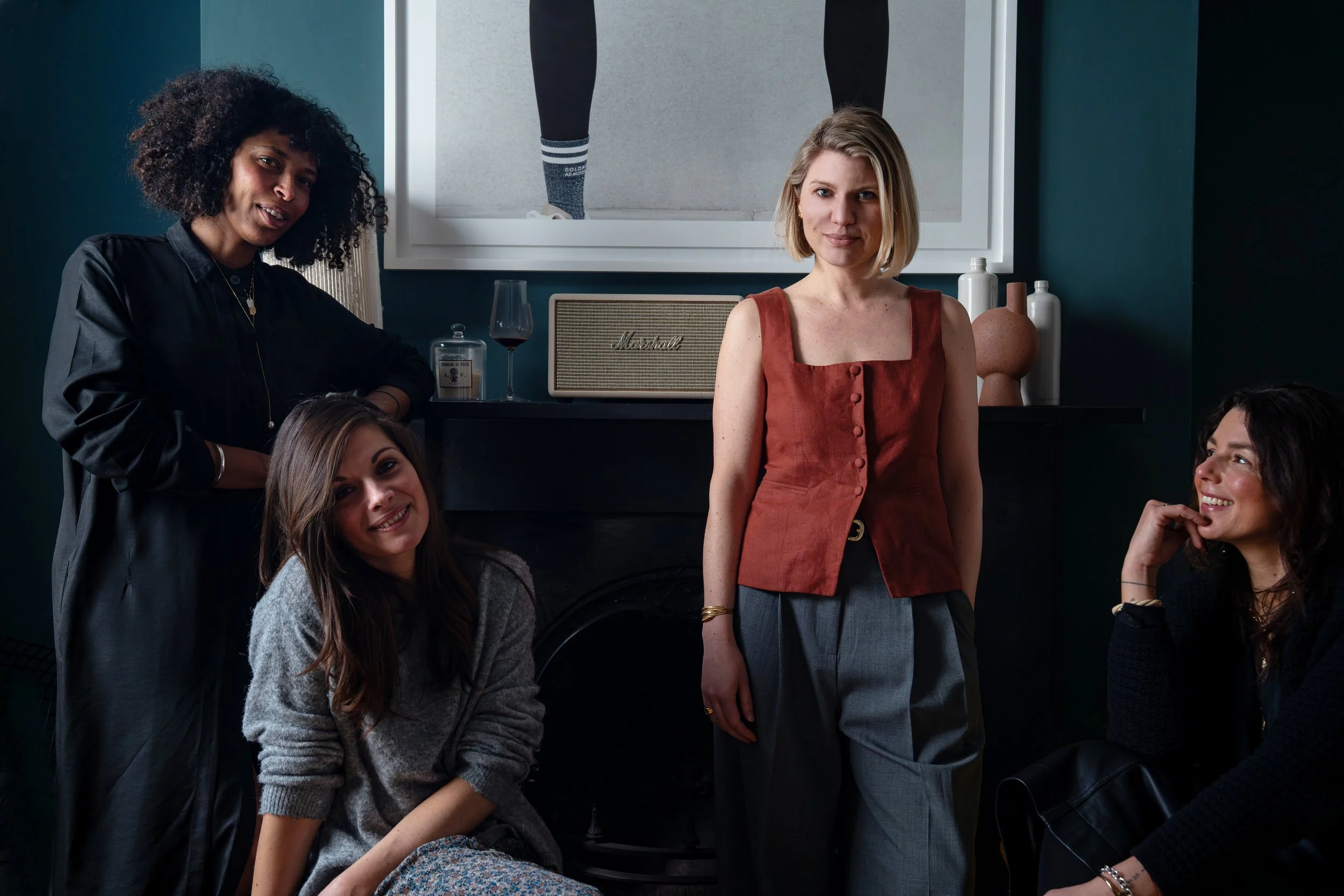 Five women gathered in a room with dark walls, some sitting and some standing. One woman in a red sleeveless top stands in the center, smiling. The others sit around her, with two women smiling and one looking thoughtful. On the fireplace mantel behind them, there is a glass of red wine, a candle, a marshall speaker, and decorative vases, with a large framed artwork displaying a person's legs in the background.