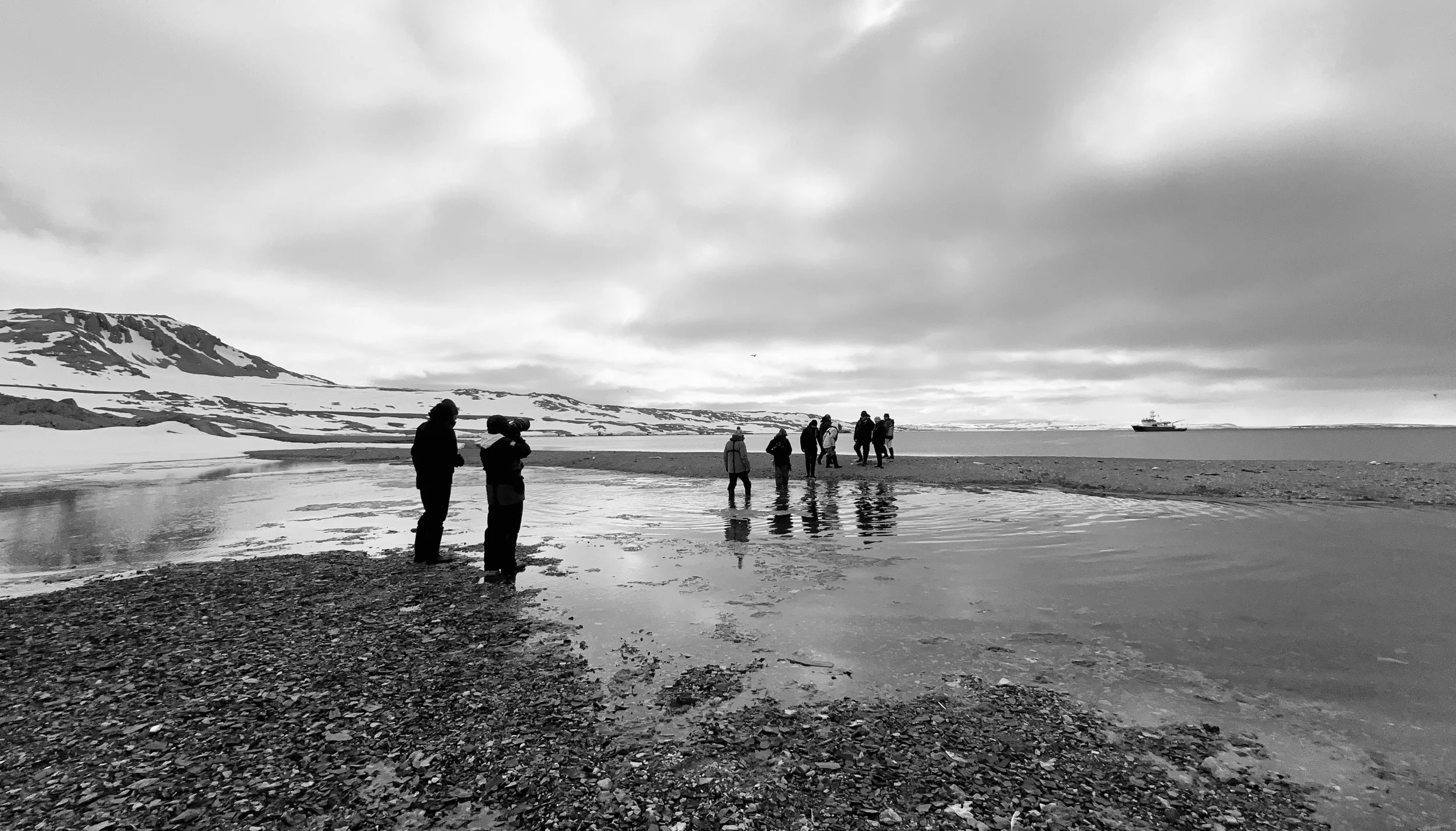 A group of people standing and walking along a pebble beach near water, with snow-covered hills in the background and a ship visible on the horizon, under a cloudy sky.
