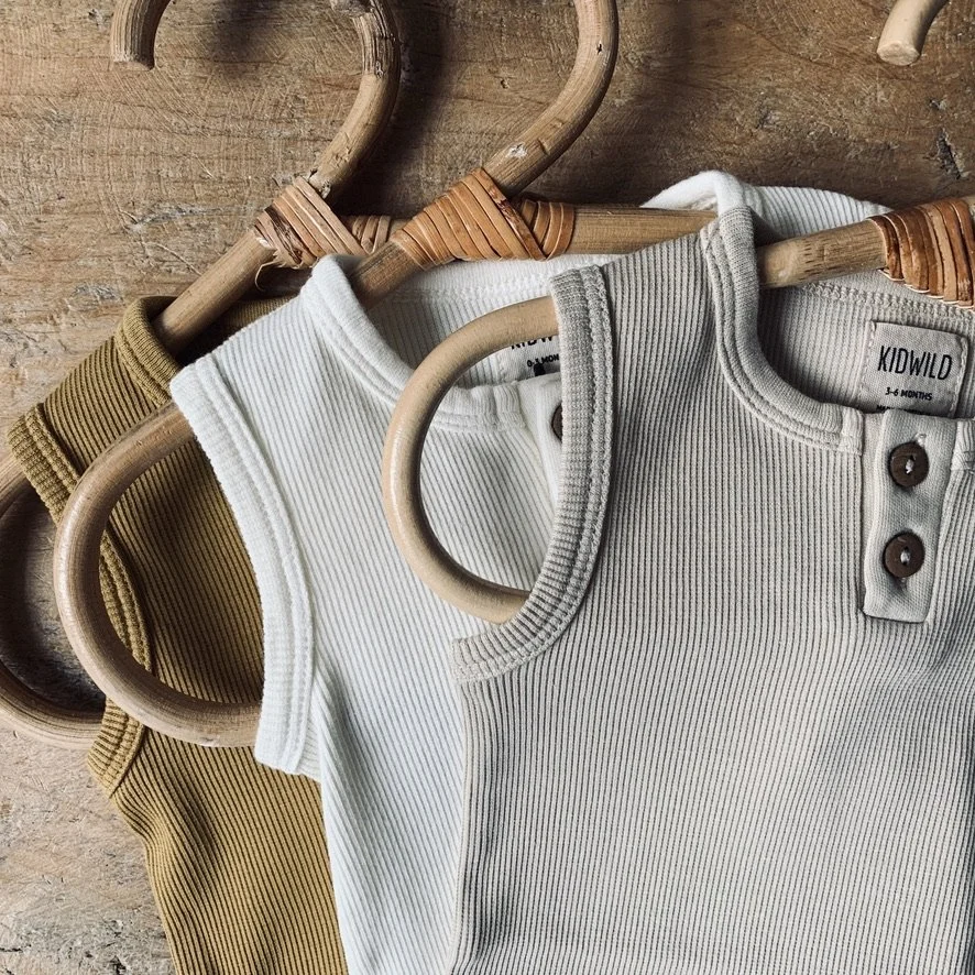 Three sleeveless baby onesies in white, tan, and mustard are hanging on wooden hangers against a wooden background.