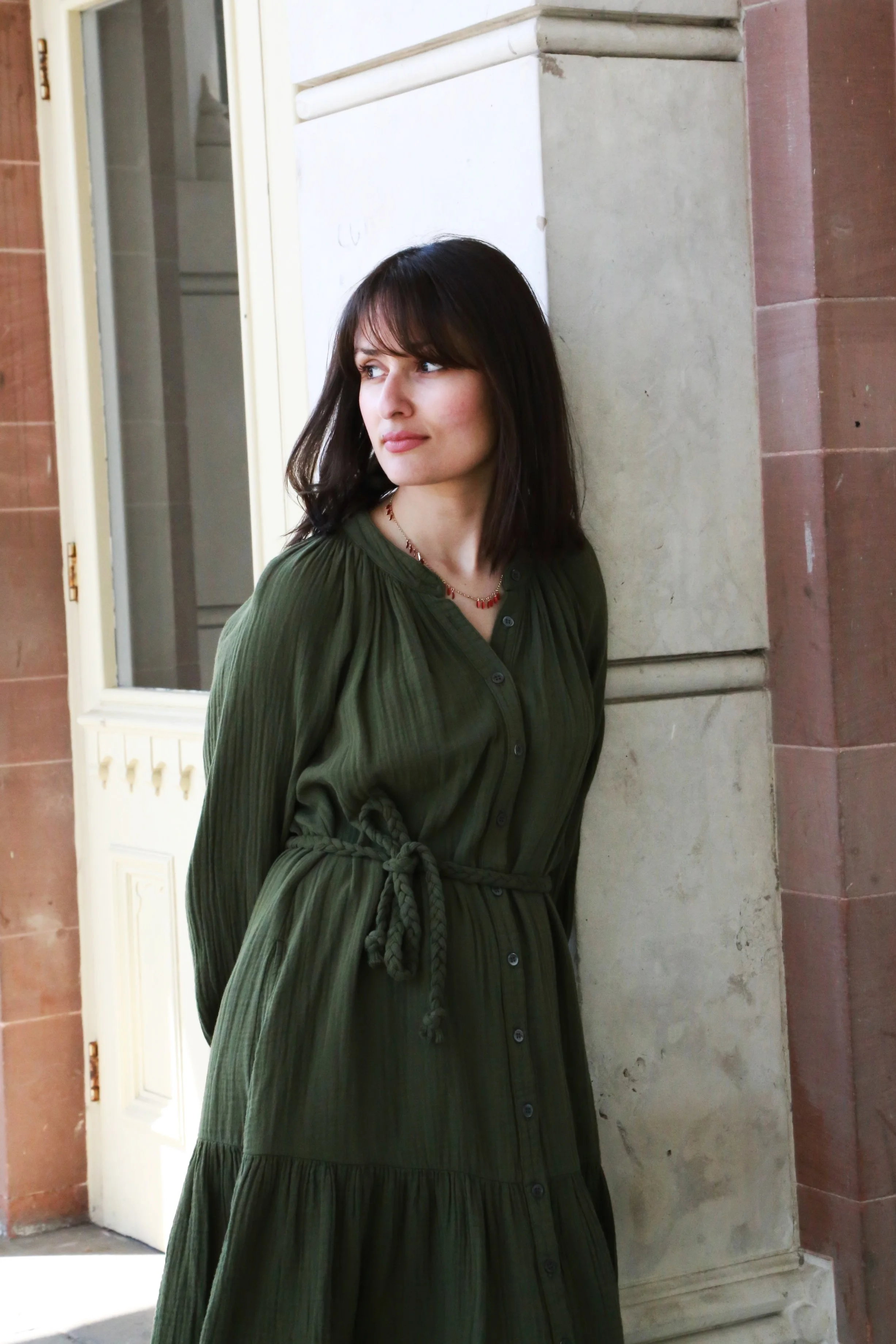 A woman with dark brown hair wearing a green dress with a waist tie, standing against a stone wall outside a building.