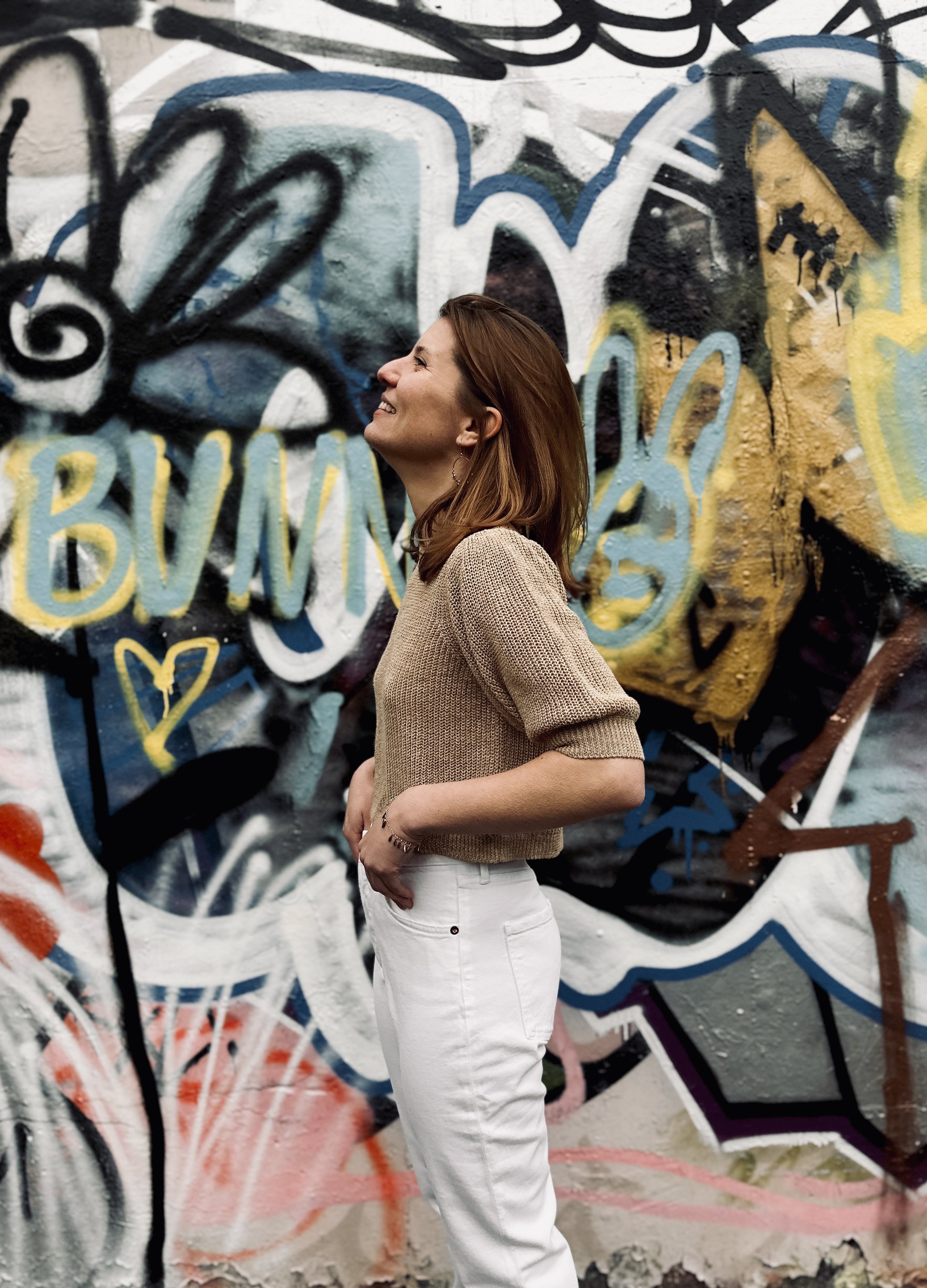 A woman with shoulder-length brown hair smiling and standing in profile against a graffiti-covered wall.