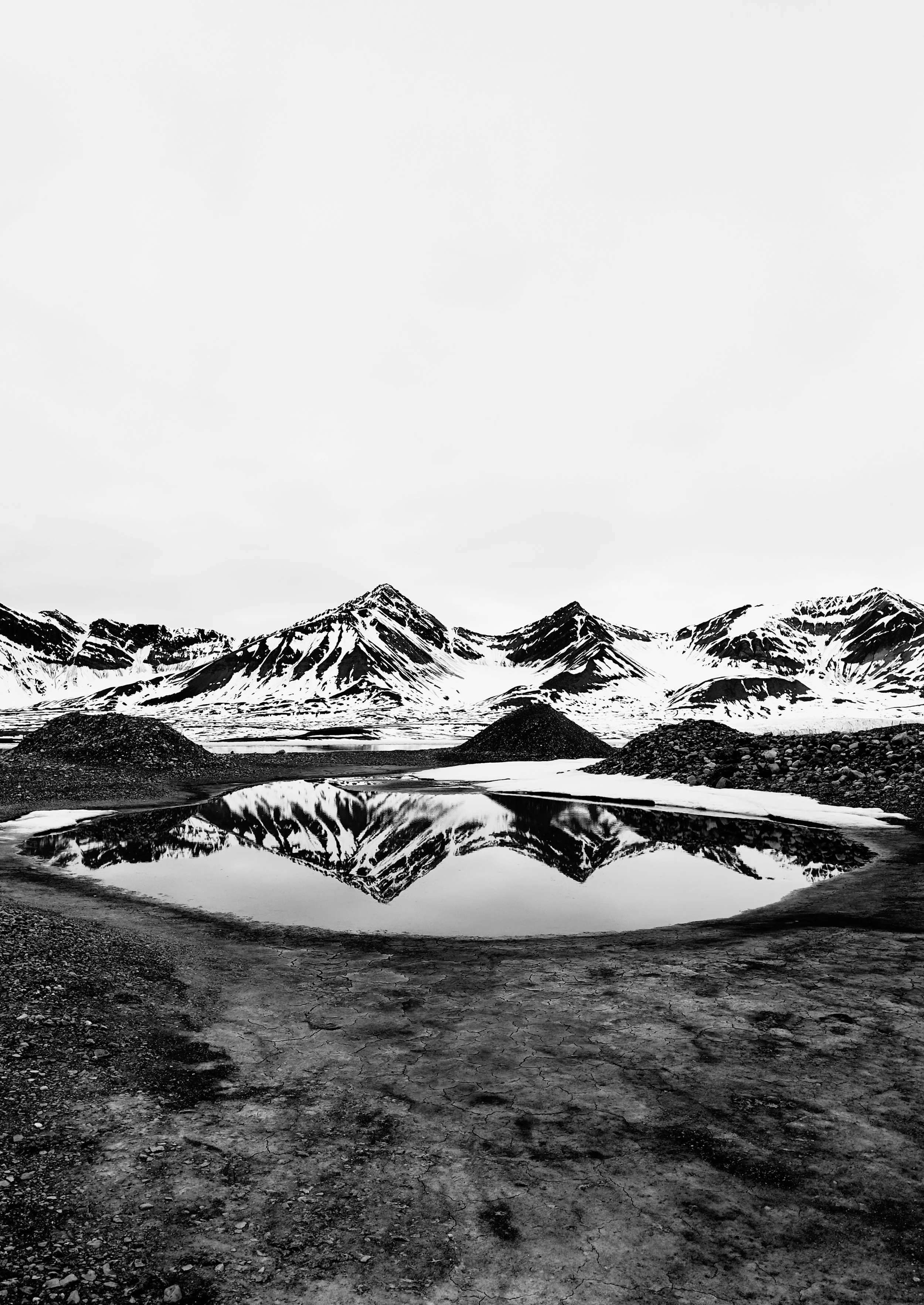 A black and white landscape of snow-capped mountains reflected in a small body of water with cracked ground in the foreground.