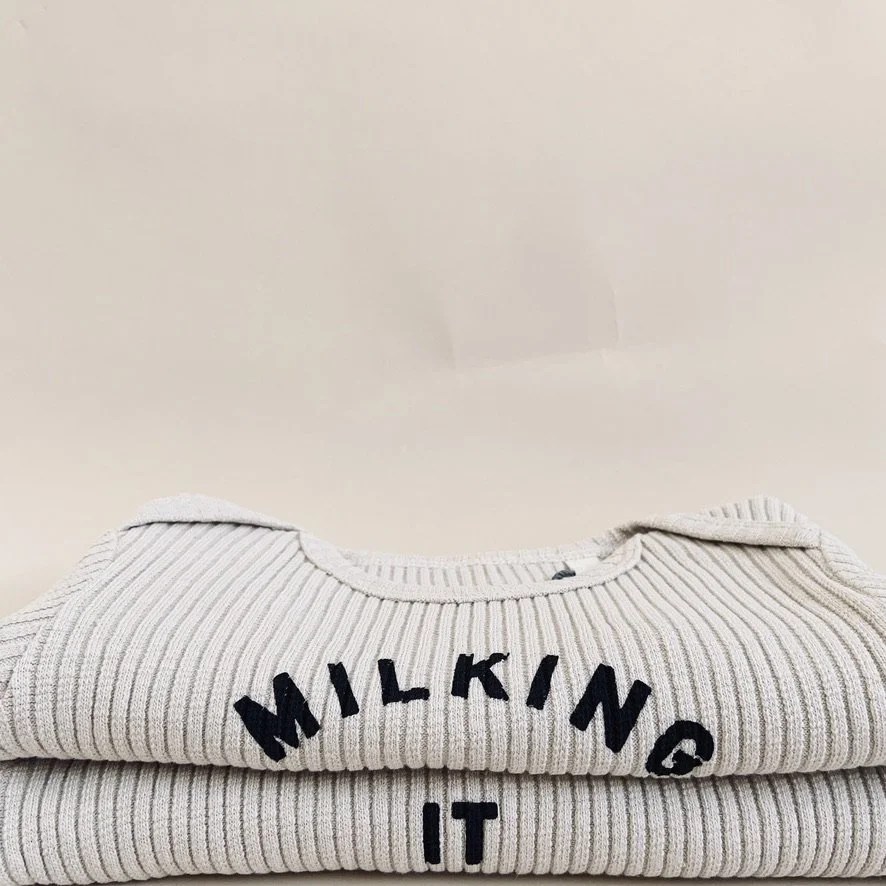 Close-up of folded beige ribbed sweater with black embroidered text "MILKING IT" in a stack.