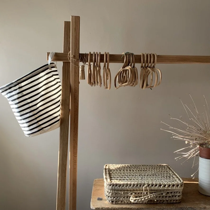 Wooden coat rack with hanging wooden hangers and a striped fabric bag, next to a woven basket on a wooden table, with a potted plant nearby.