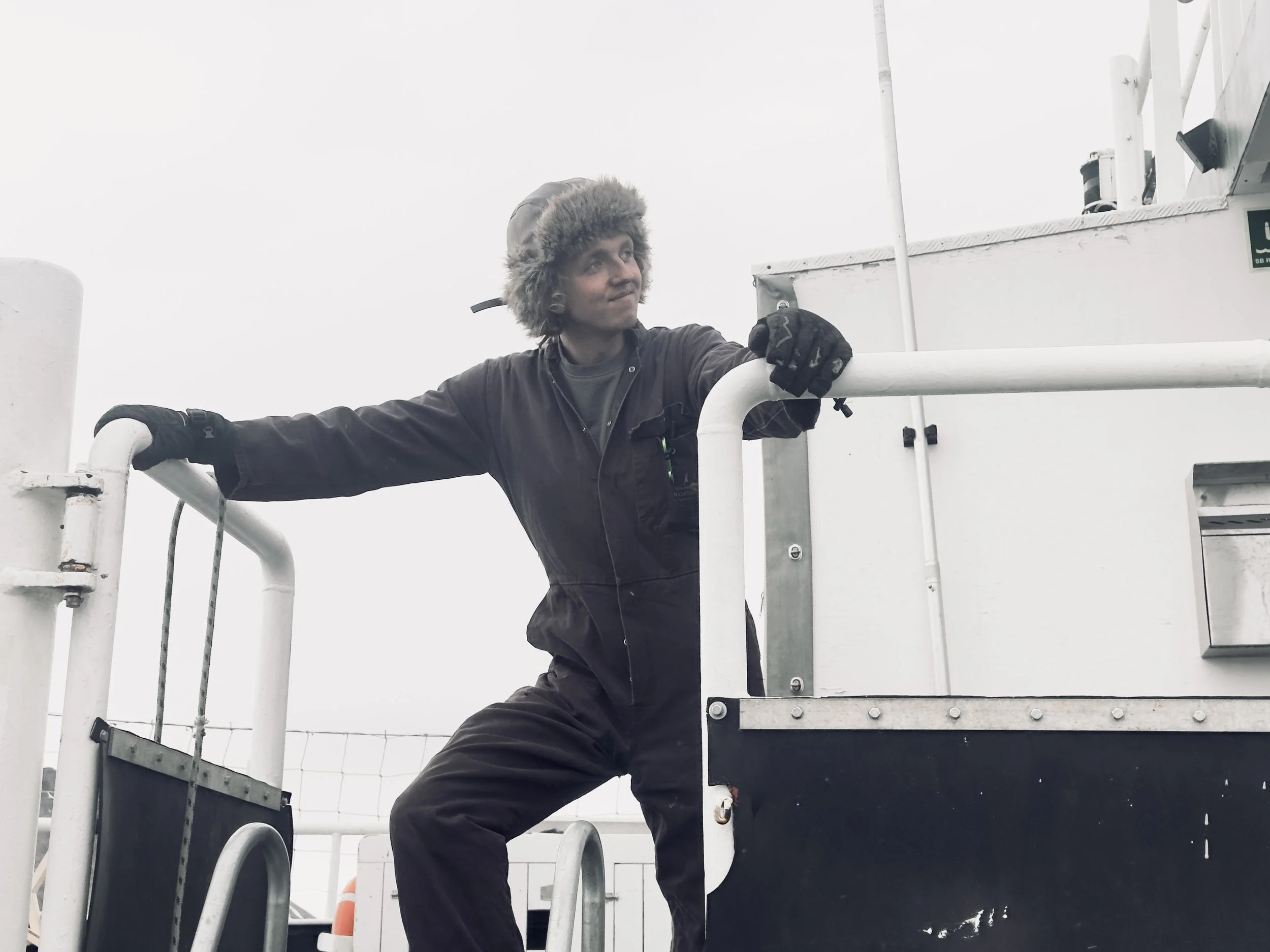 A person dressed warmly with a fur-lined hat and gloves, standing on a metal platform of a ship or industrial structure, holding onto a white railing.